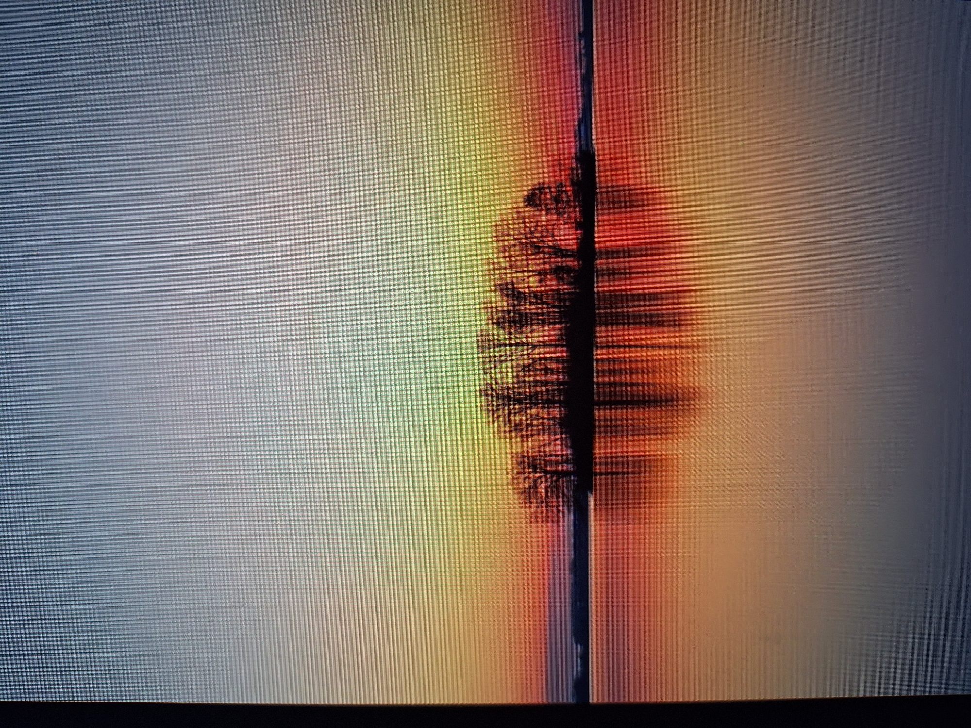 This image depicts a serene sunset over a tranquil body of water, possibly a lake or a calm sea. A small, silhouetted island covered with bare trees stands in the middle distance. The sky transitions through a gradient of colors, from a soft blue at the top to warm hues of orange, red, and yellow near the horizon, creating a beautiful reflection on the water's surface. The scene is devoid of any people or explicit man-made structures, evoking a sense of natural tranquility. The image does not contain any visual information that would allow for its localization to the city of Varna, Bulgaria.
