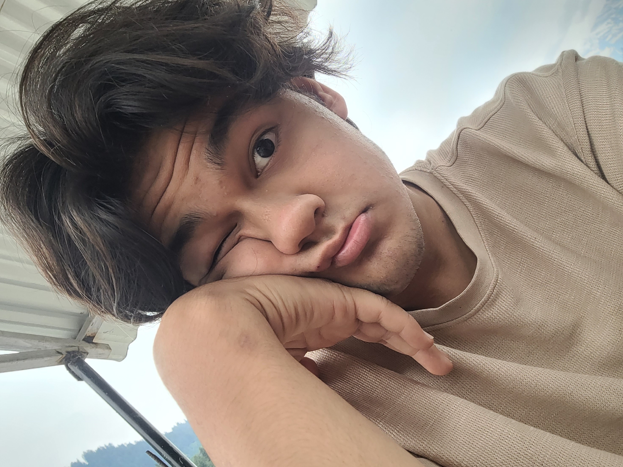 A young male is pictured from the chest up, outdoors in Vashisht, India. He is leaning his left hand against his cheek and making a pouty expression. He has dark, messy hair, brown eyes, and is wearing a beige crewneck shirt. The background consists of a cloudy sky and hints of green foliage.