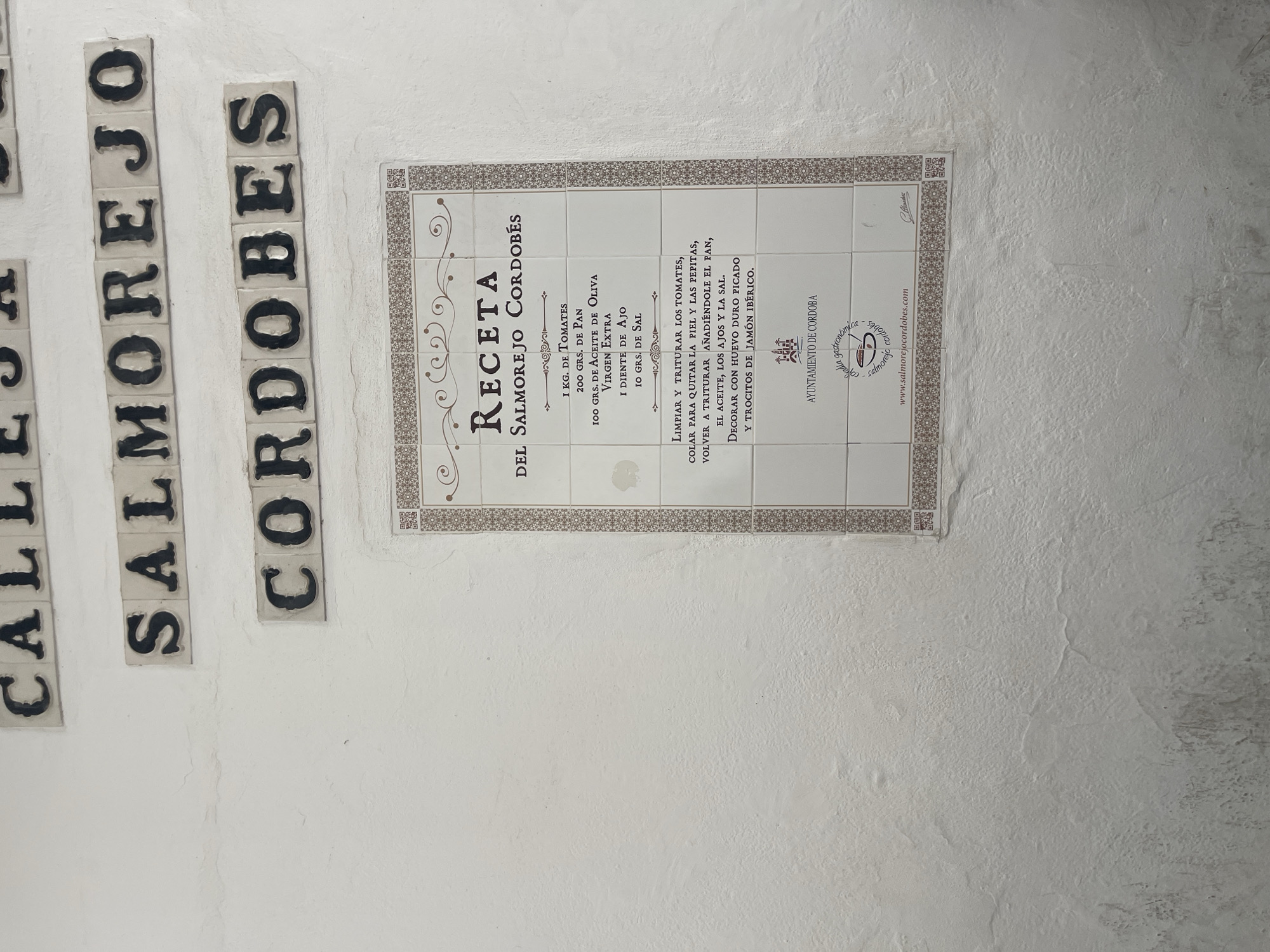 The image shows a tiled plaque on a white stucco wall, located in Córdoba, Spain. The plaque displays a recipe for "Salmorejo Cordobés" and includes text in Spanish. Above the plaque, there are other tiled inscriptions with words like "CALLE" and "SALMOREJO CORDOBES". The recipe lists ingredients such as tomatoes, olive oil, garlic, and salt, and provides instructions for preparation. Below the recipe, there is an emblem with "AYUNTAMIENTO DE CORDOBA" written on it, along with a website address "www.salmorejocordobes.com". The overall scene is an outdoor setting on a wall, likely in a public area.