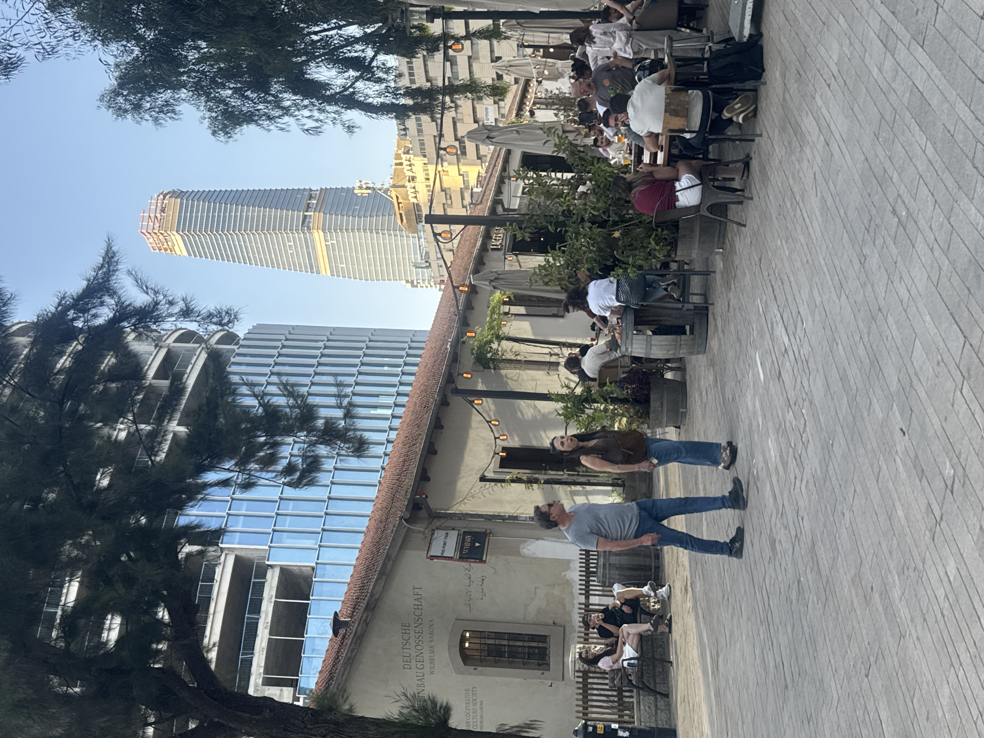 This image captures a vibrant outdoor scene in Tel Aviv, Israel, likely within the historic Sarona complex, showing a blend of old and new architecture. The setting features a wide pedestrian walkway paved with light-colored stones, bordered on the right by a historic-looking building housing an outdoor cafe or restaurant.
Numerous people are visible, indicating a lively atmosphere. Many individuals are seated at tables under large umbrellas along the right side of the walkway, engaged in dining, drinking, and socializing. In the foreground, a man and a woman are walking along the paved path. Further down, other people are seen relaxing on benches outside the building.
The historic building itself has a tiled roof and traditional windows. Beyond the immediate cafe area, modern high-rise buildings dominate the background, with one particularly tall skyscraper appearing to be under construction, showcasing a contemporary facade. Green foliage from trees is interspersed throughout the scene, providing shade and natural elements.
The lighting suggests late afternoon or early evening on a clear, sunny day, with soft shadows cast across the pavement. Visible text on the historic building identifies it as the "DEUTSCHE EINBAU GENOSSENSCHAFT" (German Construction Cooperative) and "WILHELMA SARONA," with translations also in English and Arabic, reinforcing the historical German Templer colony context of Sarona. Other signs include "FIVE POINT FOUR," "LAGER&," "WHIST" (likely "WHISKY"), and "שטחים ל" (Hebrew for "areas for"). The image conveys a sense of urban leisure and cultural heritage.