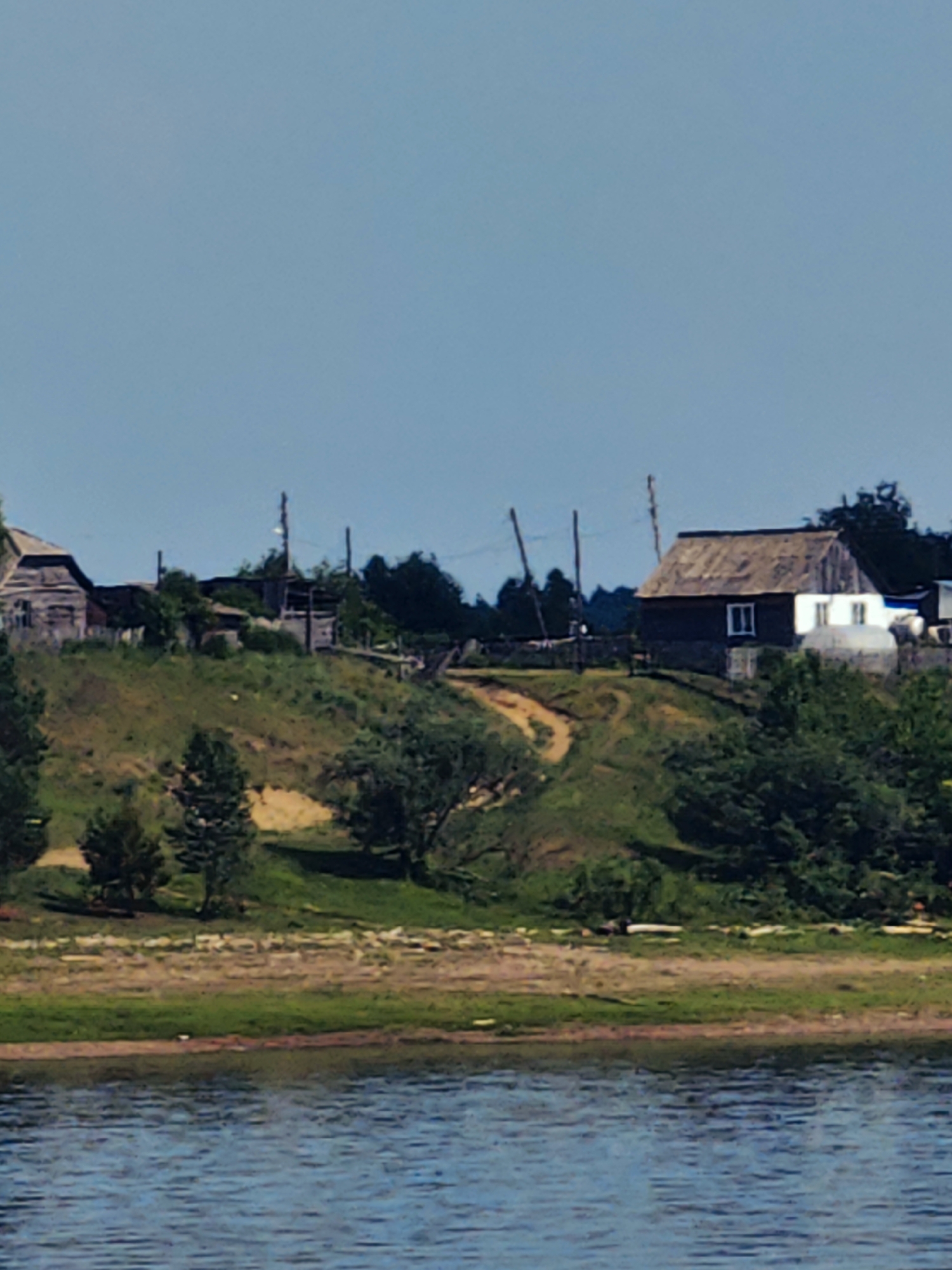 The image shows a pastoral scene with a village nestled on a grassy hill overlooking a body of water. The sky is clear and blue. On the hill, there are several wooden houses, some appearing weathered and old, with visible roofs and windows. A dirt path winds its way up the hill. Green trees and foliage are scattered throughout the landscape, with a denser cluster of trees on the right side of the hill. The foreground features the tranquil blue water. The scene evokes a sense of rural tranquility.