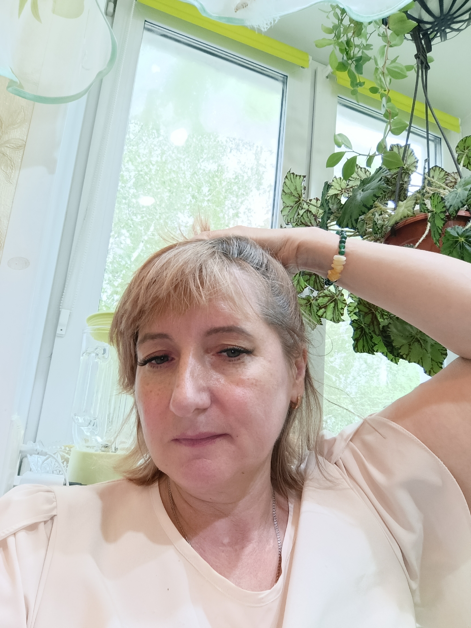 The image shows a medium shot of a middle-aged Caucasian woman indoors, possibly at home in Tugulym, Russia. She is looking at the camera with a neutral expression, her hand resting on her head. She is wearing a light pink, loose-fitting top and a beaded bracelet on her wrist. The background features a window with light filtering through, suggesting it is daytime. Green foliage from a hanging plant is visible on the right side of the image. Other objects, like a glass pitcher, are partially visible in the background. The overall impression is of a casual, everyday moment.