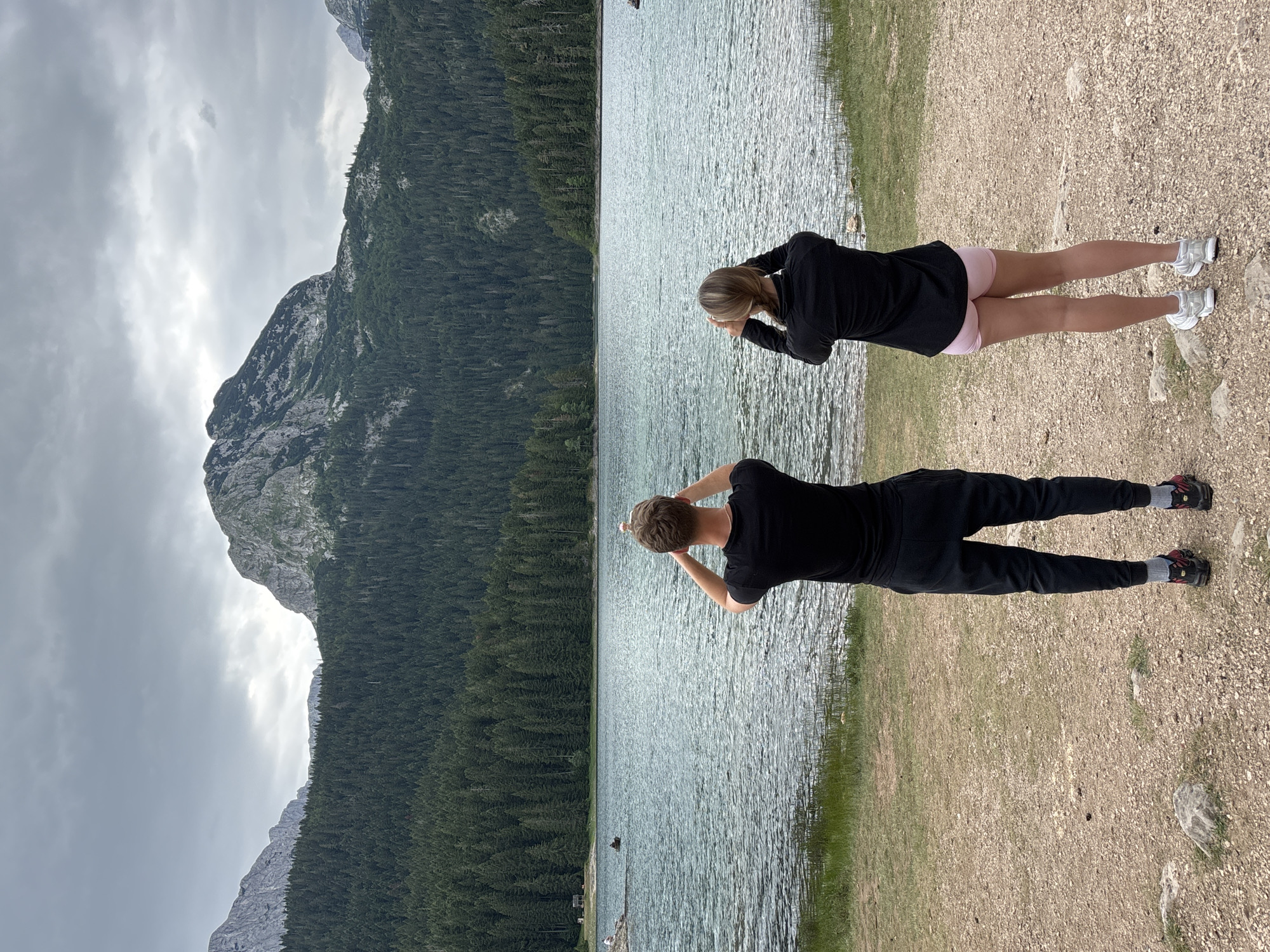 In Pitomine, Montenegro, a man and a woman stand facing away from the camera, looking out over a large, calm lake. The man, on the left, wears a black t-shirt and black pants, with red and black athletic shoes. The woman, on the right, wears a black jacket and pink shorts, with white athletic shoes. Both are holding their hands up to their faces, as if looking through binoculars or taking photographs.
The lake's surface is a translucent blue-green, with gentle ripples. The shoreline in the foreground consists of sandy and gravelly ground with sparse green grass. Behind the lake, a dense, dark green forest of pine trees covers the sloping hillsides. Towering over the trees in the distance are rugged, rocky mountains under a cloudy, overcast sky. The overall atmosphere is one of serene natural beauty.