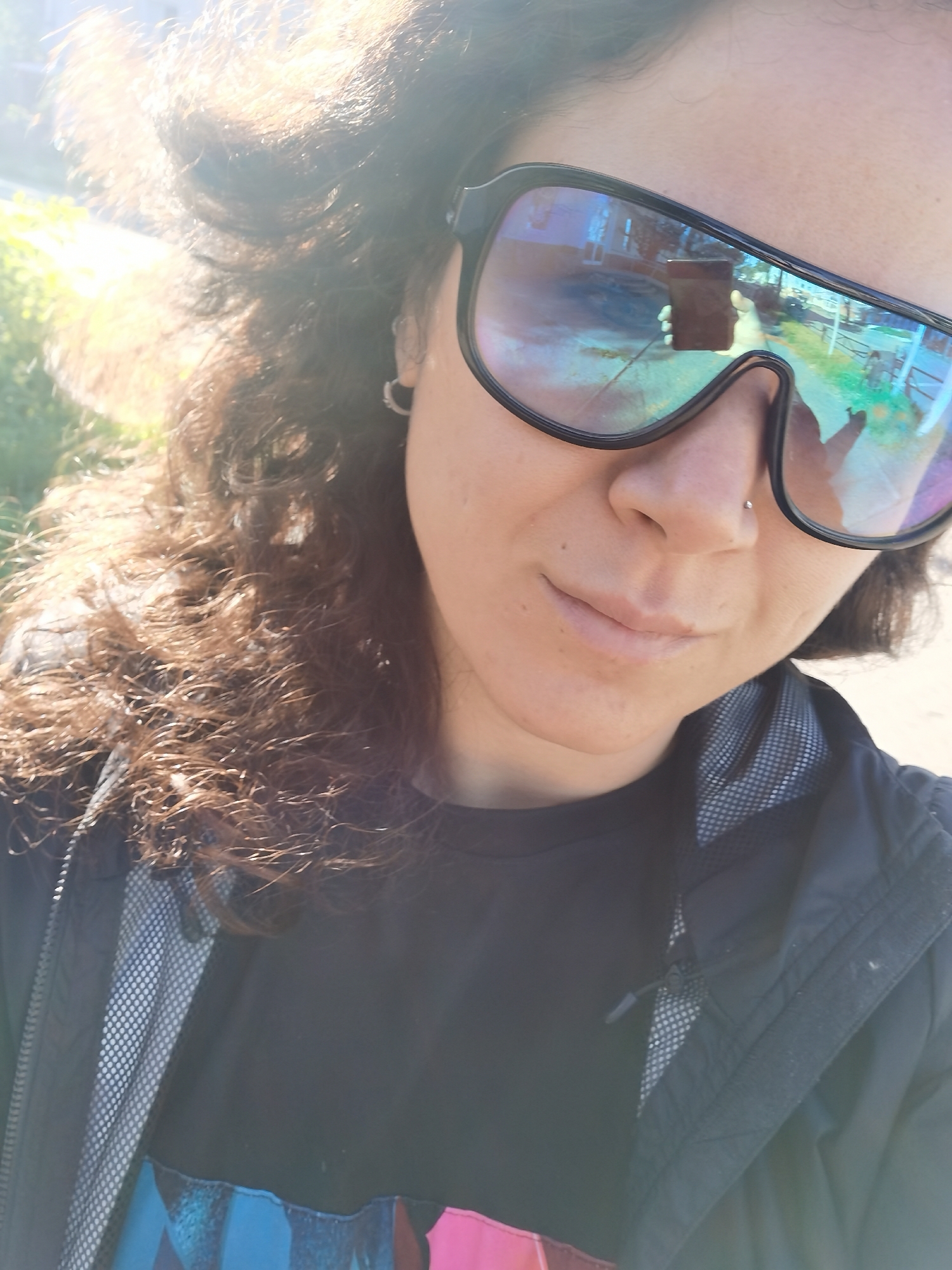 A woman with curly brown hair and a nose ring is taking a selfie. She is wearing large, reflective sunglasses and a black t-shirt under a dark jacket with a grey mesh lining. The sunglasses reflect a street scene with houses and trees, and a hand holding a phone. The image appears to have been taken outdoors in Chaikovsky, Russia, as evidenced by the architectural style of the reflected buildings and the presence of birch trees, common in the region. The lighting suggests it is daytime with bright sunlight.