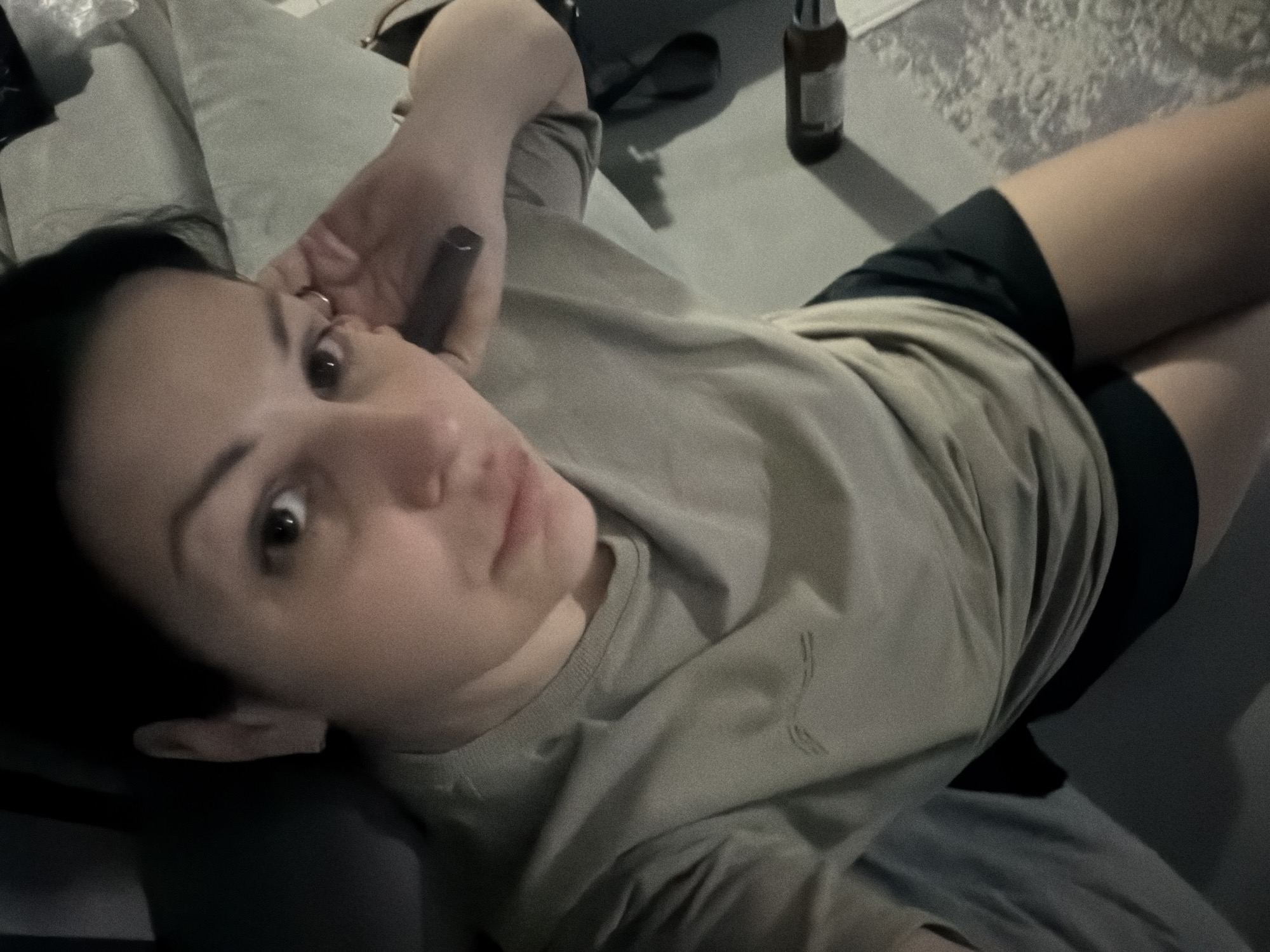 A woman is lying down on a sofa, wearing a light olive green t-shirt and black shorts. She is looking up towards the camera, with her right hand near her ear and holding a dark object, possibly a vape pen. Her left arm is bent, with her hand resting on her chest. The background is dimly lit, with a textured rug visible on the right side of the image and what appears to be a bottle on the rug. The scene is indoors, in what could be a living room. There are no visible landmarks or indicators that would place this scene specifically in Sochi, Russia.