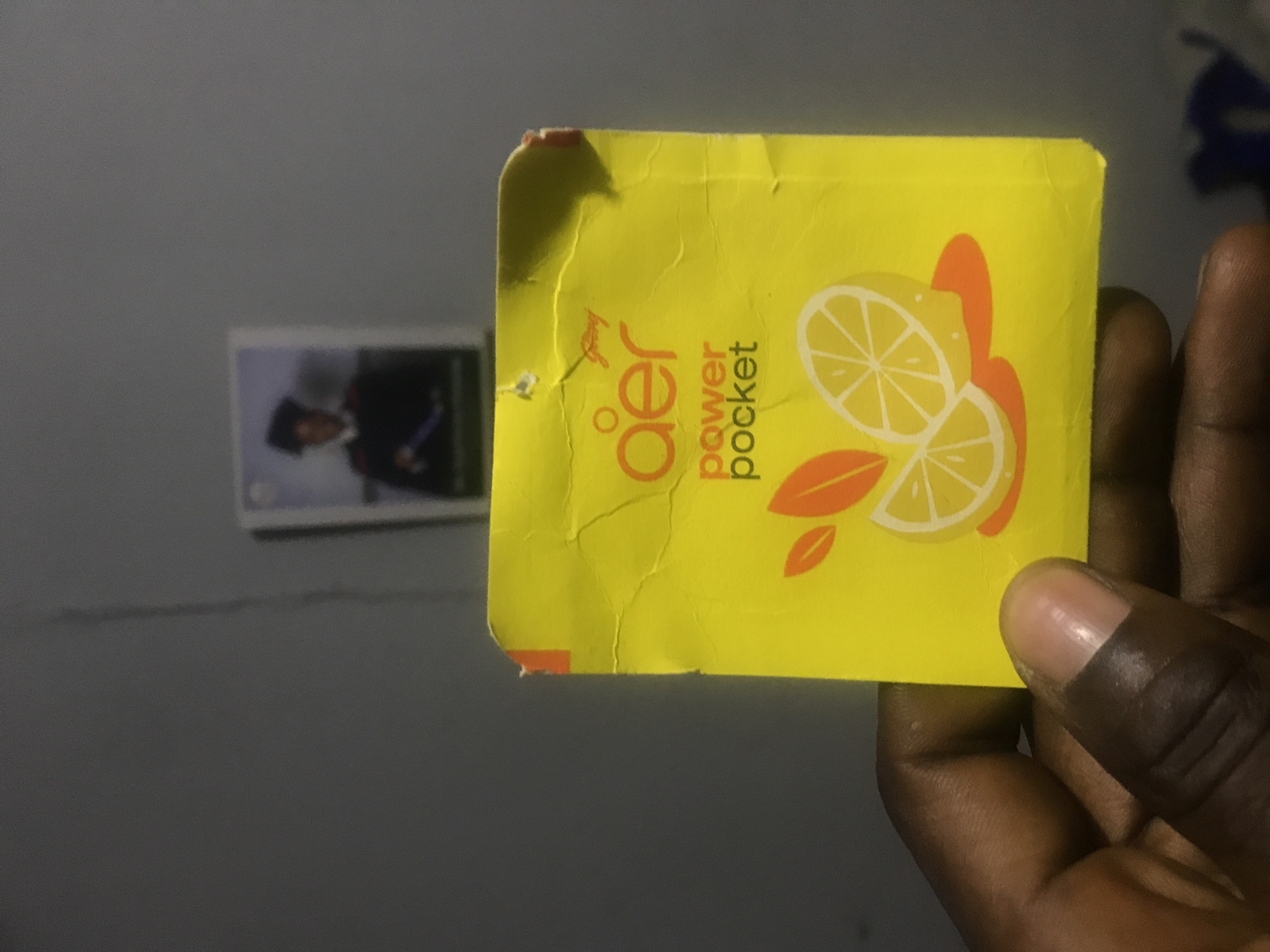 A dark-skinned hand holds a yellow rectangular packet, possibly an air freshener, in a close-up, vertically oriented shot. The packet is slightly creased and features the brand name "Godrej" in small text at the top, along with prominent text reading "aer" and "power pocket." An illustration of sliced lemons and orange leaves is also visible on the packet's surface. In the blurry background, a framed picture hangs on a plain gray wall, depicting a person in what appears to be formal attire or a uniform. The indoor lighting suggests artificial illumination, casting subtle shadows. The scene captures a moment of someone holding or presenting the product. This image was taken in Rafin Par, Nigeria.