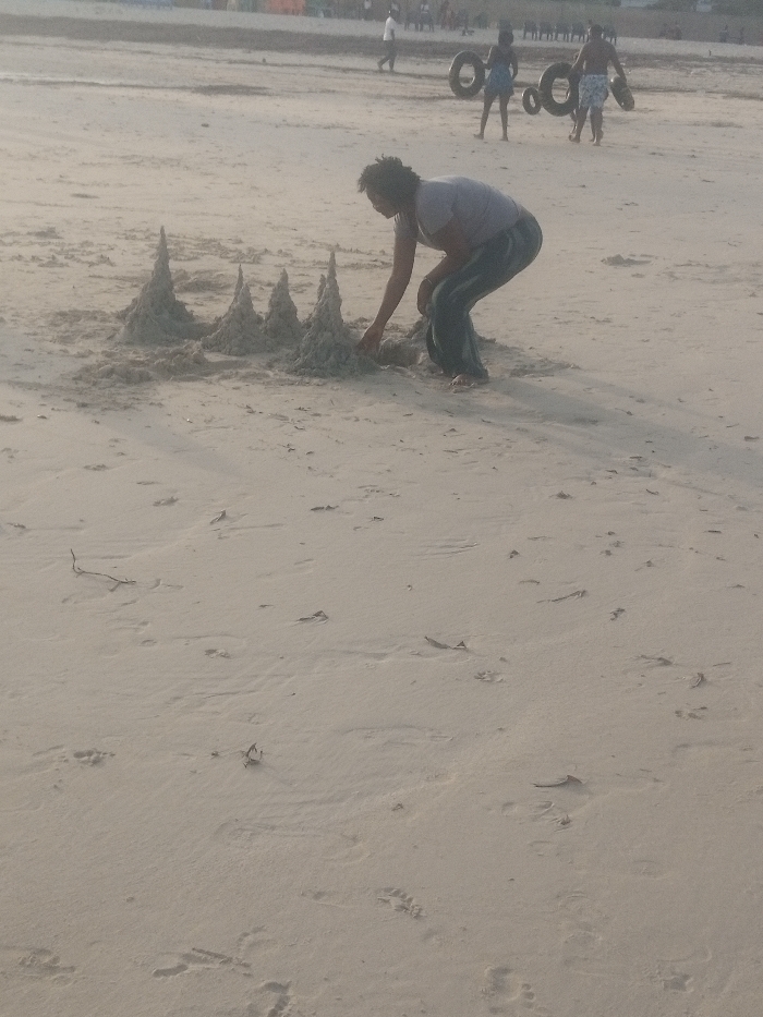 The image depicts a daytime scene on a sandy beach in Shanzu ward, Kenya. In the central foreground, an individual with dark, curly hair, dressed in a light-colored short-sleeved top and dark trousers, is crouched down, actively shaping a sand structure with their hands. This structure consists of five distinct, conical sand mounds arranged in a row. The individual is barefoot. In the mid-background, several other people are present on the beach; one figure is walking to the left, while two figures further right are carrying large, dark, circular objects resembling inner tubes or tires. The sandy surface is light-colored, showing numerous footprints and scattered small pieces of organic debris. The scene captures beach recreation.