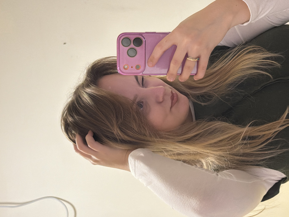 A young woman takes a selfie. Her blonde and brown hair is partially obscuring her face. She is wearing a white long-sleeve shirt under a dark-colored vest. She holds a pink phone with three camera lenses. A gold ring is visible on her finger. One hand is touching her hair, near her forehead. A light-colored wall is visible in the background, with a small dark spot near the top. A white cable is partially visible in the lower left corner.