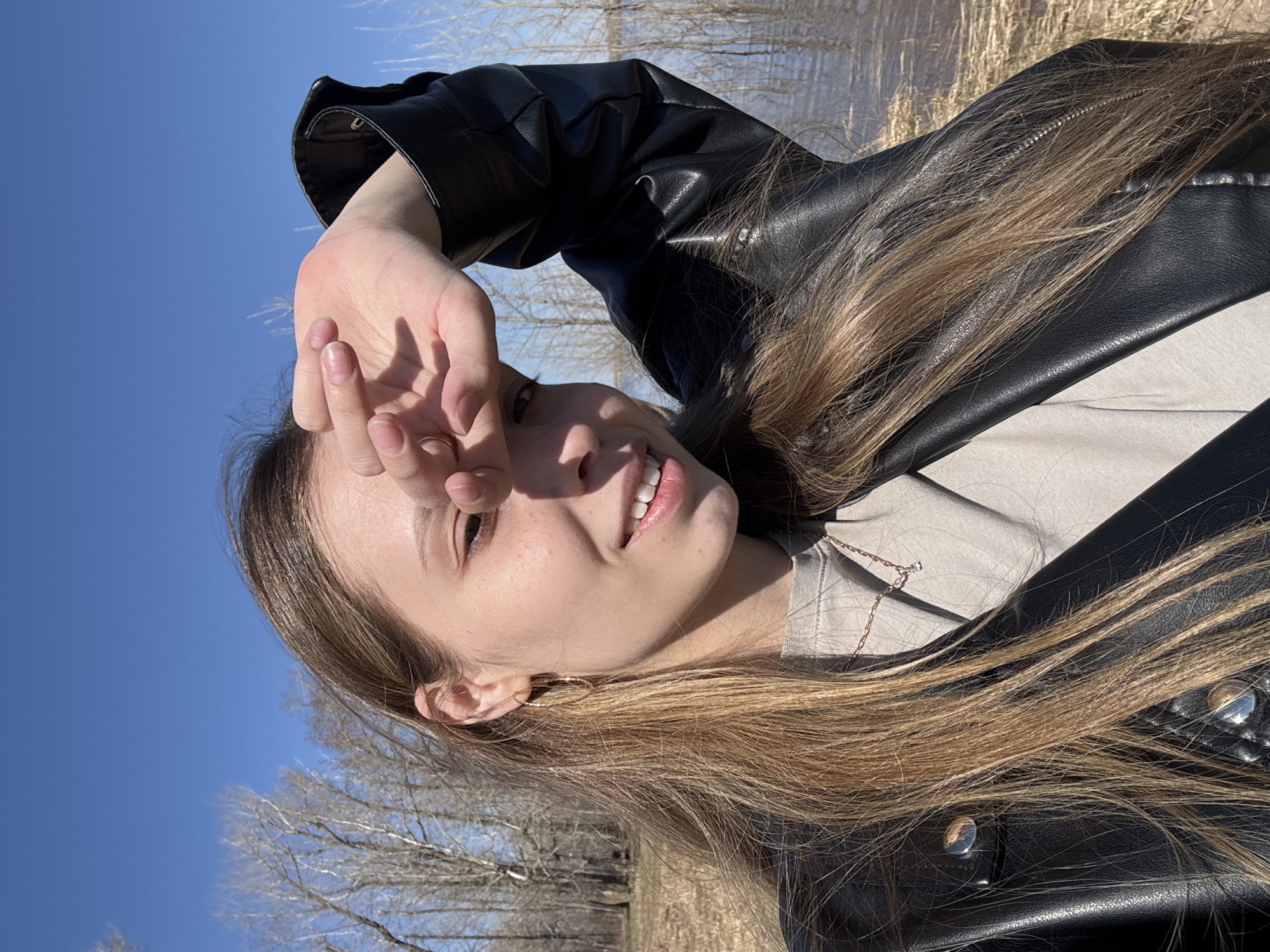 A young woman with long, light brown hair poses for a selfie outdoors on a sunny day. She is wearing a black leather jacket and a light-colored shirt underneath, with a delicate gold chain necklace visible. She holds her hand up to shield her eyes from the sun, smiling slightly. The background consists of a blue sky and bare trees, suggesting a location in a park or near a river. Given the prompt's mention of the city of Chaikovsky, Russia, it is plausible the image was taken in a park or natural area within or near that city, near a river that is visible in the image.