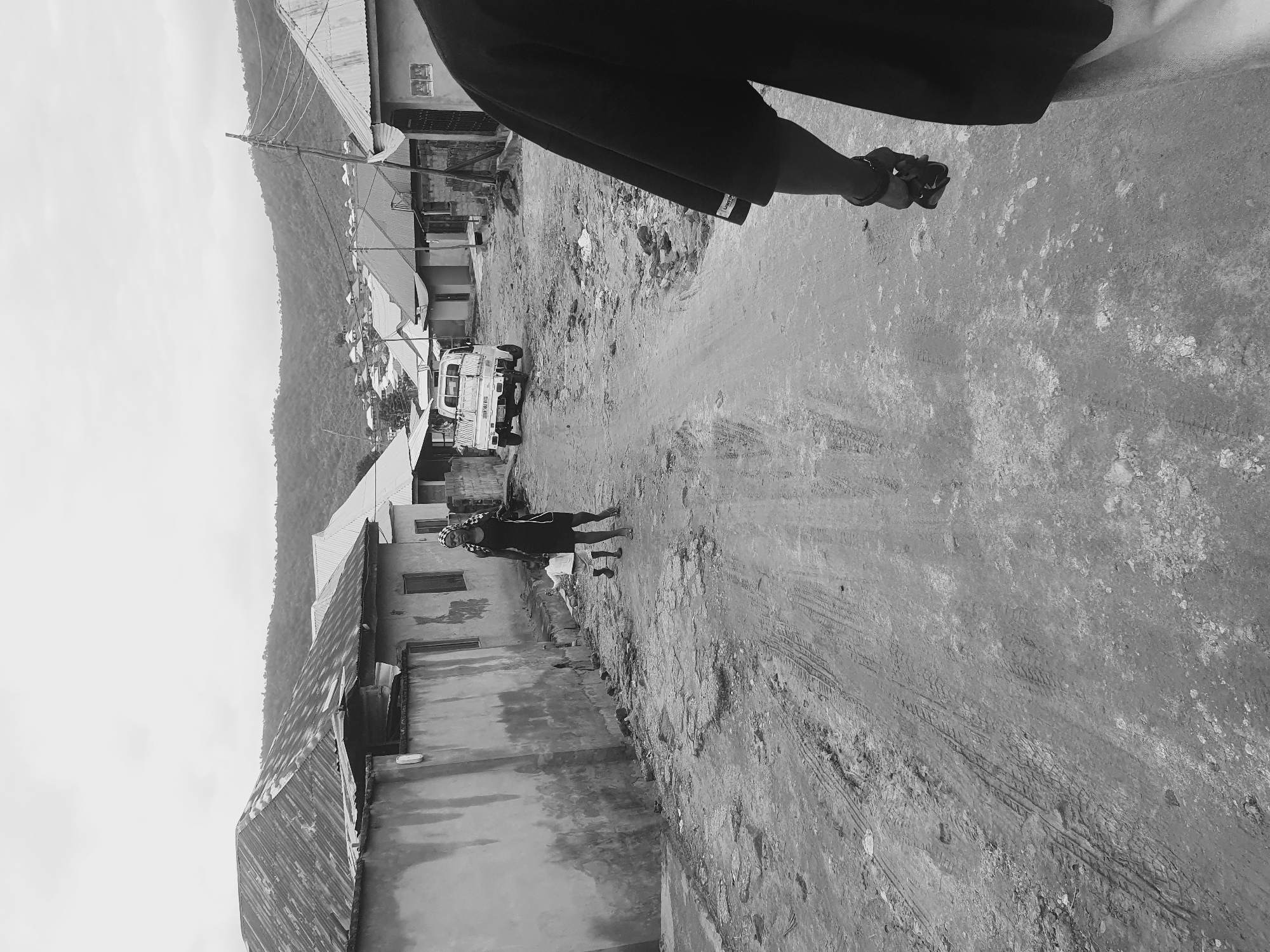 The image depicts a street scene in Lokoja, Nigeria. The road is unpaved, with visible tire tracks and debris. A white van with visible text on its side is parked on the road. A person is walking on the road. Buildings with corrugated metal roofs line the street. Hills are visible in the background. A person wearing sandals and black pants is partially visible in the top right of the image.