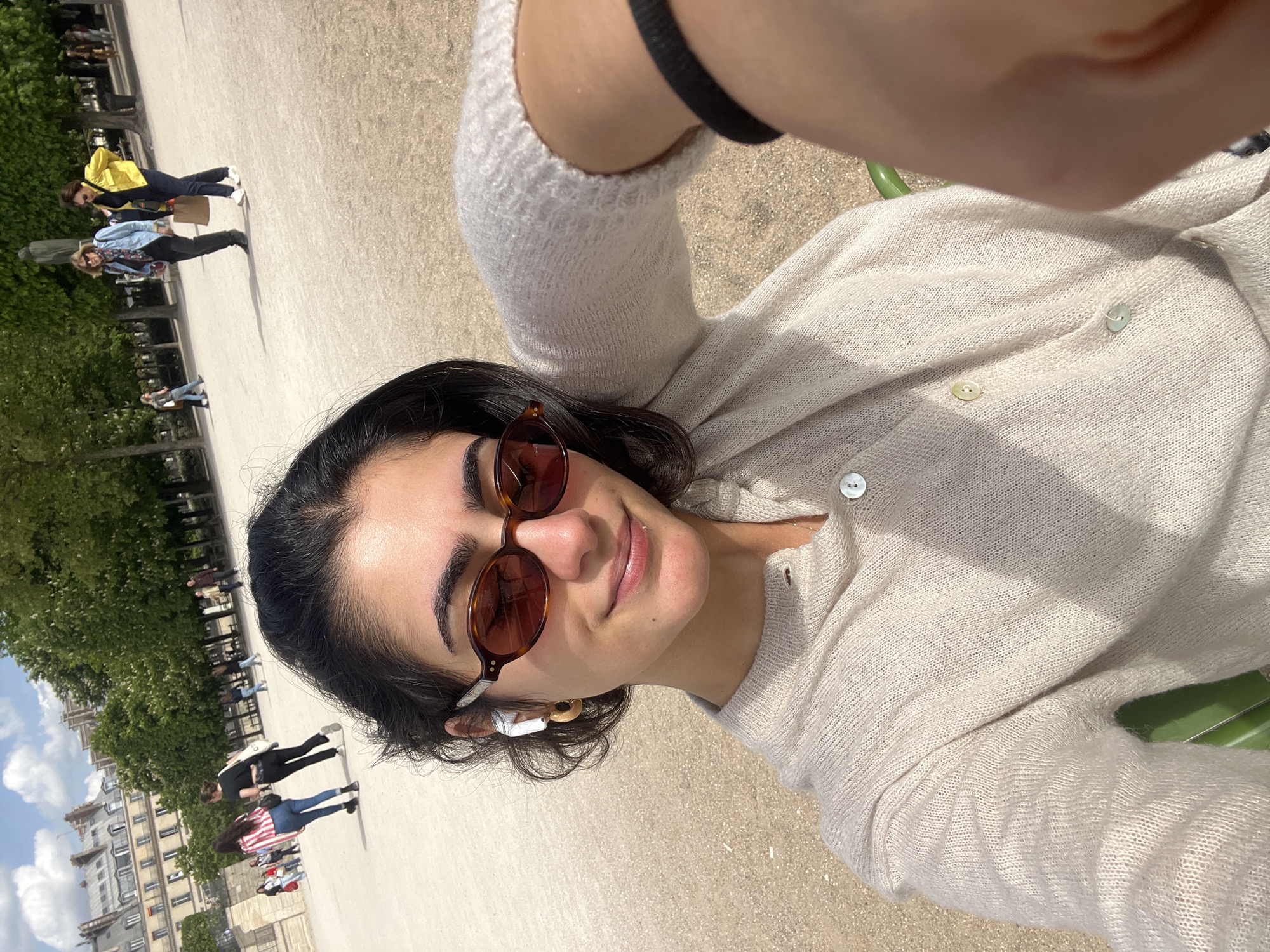 The image is a bright, sunny outdoor selfie-style portrait, rotated 90 degrees clockwise. The main subject is a young woman, positioned in the foreground and smiling slightly towards the camera. She has dark hair and is wearing brown-framed sunglasses and a light beige or off-white, softly knitted cardigan with multiple light-colored buttons. A white earbud is visible in her left ear, along with a gold-toned earring. Her arm, partially visible, appears to be holding the camera.
The background shows a wide, open public space, likely a park or large promenade, with a light-colored ground surface. Green trees are prominent, and in the distance, various people are seen walking, some casting long shadows, suggesting it is midday or early afternoon. Buildings with classic European architectural styles are visible further in the background, consistent with a Parisian setting. No visible text is present in the image. The overall scene conveys a pleasant, relaxed atmosphere.