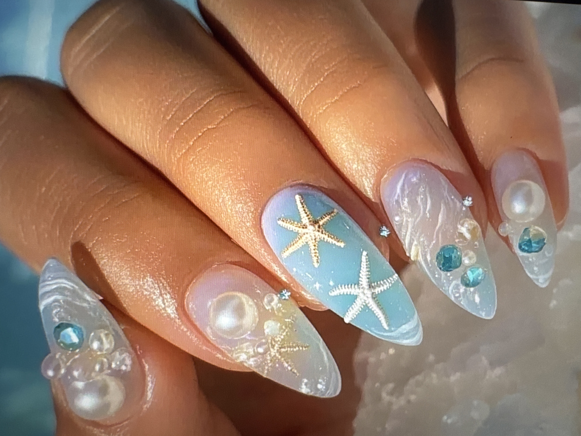 A close-up shot features a person's hand with elaborate nail art. The nails are long and stiletto-shaped, decorated with a beach and ocean theme. Some nails are adorned with pearls, small iridescent beads, and blue rhinestones, mimicking seafoam and water droplets. Others feature miniature starfish charms, one in gold and two in silver, placed on a light blue background that transitions to a pale purple at the cuticle, suggestive of a sunset over the ocean. The skin of the hand appears to have a healthy, warm tone. The background is blurred, but suggests a bright, possibly outdoor setting with a hint of turquoise, possibly a sky or water. There is no identifiable location within the city of Tallinn, Estonia.