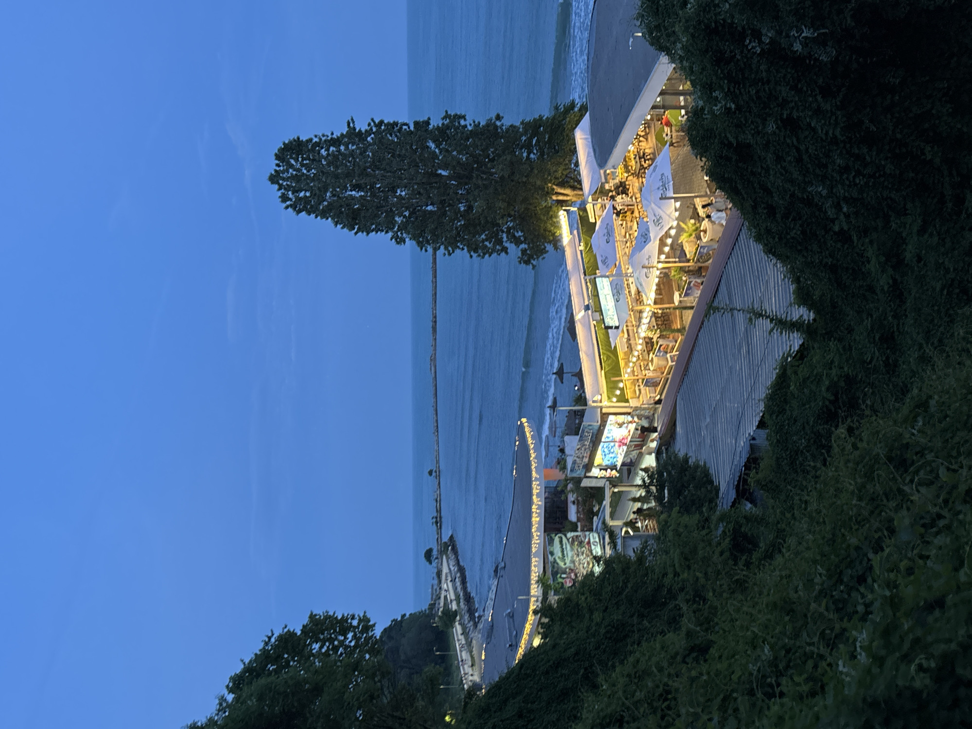 This image captures a vibrant coastal scene at dusk or early night in what appears to be Varna, Bulgaria. The sky is a deep, clear blue with faint wisps of clouds, indicating the transition from day to night.
In the foreground and to the right, lush green foliage and parts of a building with a corrugated metal roof frame the scene. The main subject is an inviting outdoor restaurant or bar situated along the coastline. This establishment is brightly illuminated by numerous warm string lights and interior lighting, creating a lively atmosphere. Many white umbrellas, some clearly displaying the "Kamenitza" logo, are spread across the dining area, sheltering tables where people are visibly seated, enjoying meals or drinks. The presence of patrons suggests an active dining or social event.
A tall, dense tree stands prominently near the restaurant, providing a natural partition. To the left of the tree and extending into the background is the calm expanse of the sea, likely the Black Sea. Gentle waves can be seen near the shore. A long, narrow pier or breakwater stretches far into the water, hinting at maritime activity or coastal defenses. Below the restaurant, a paved promenade or pathway runs alongside the water, also illuminated by soft, golden lights. The overall setting combines natural beauty with a cheerful, bustling social environment, characteristic of a popular resort area.
