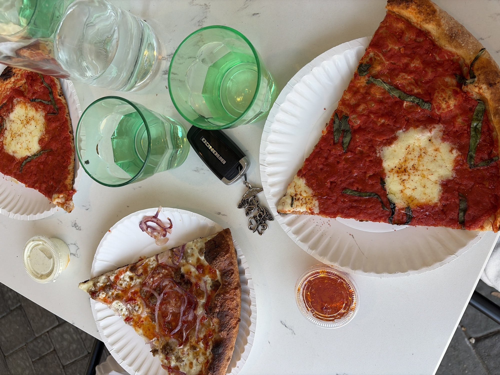 A high-angle shot shows a table with pizza slices, glasses of water, and a car key. The table surface is white, possibly marble. Three pizza slices, each on a white paper plate, are visible. One slice has a red sauce base with a central round of white cheese and scattered basil leaves. Another slice is topped with cheese, red onions, and sausage or pepperoni. A third slice resembles the first but is smaller. Three glasses of water are on the table, with two being clear and one being green. A black car key with a "BRONCO" logo is also on the table, attached to a keychain with three small butterfly charms. Two small plastic containers, one with a pale sauce and the other with a red sauce, are also present. The setting is likely a restaurant or outdoor dining area in the city of Austin, United States of America.