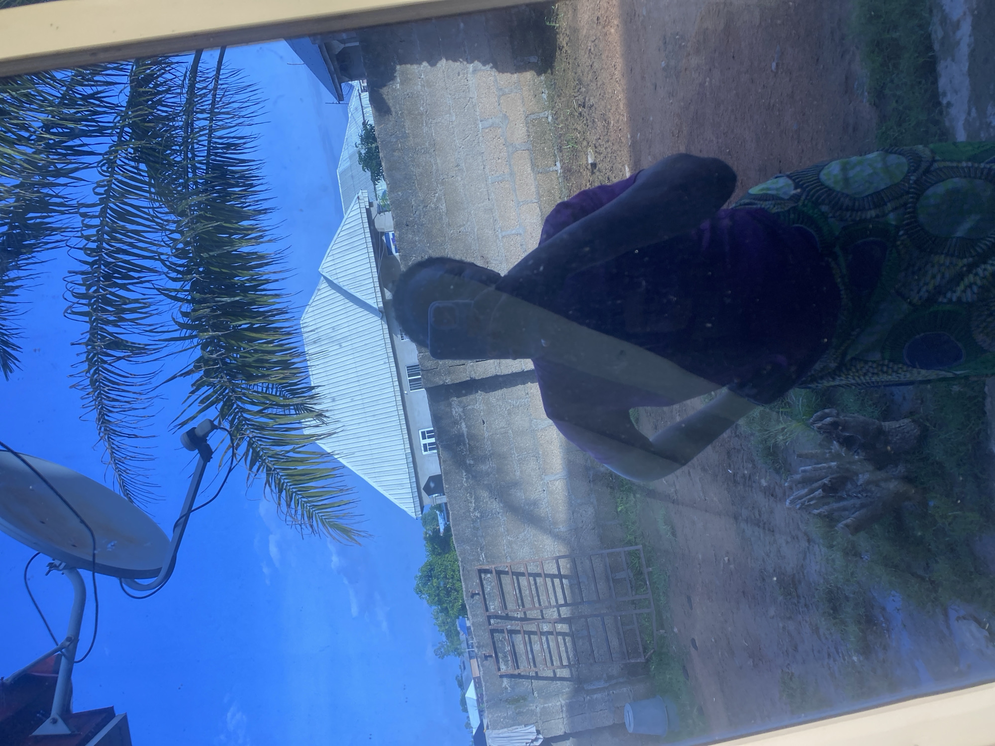 This image, taken in Jalingo, Nigeria, appears to be a reflection on a window. The primary subject is a person seen in reflection, wearing a purple top and a patterned skirt with green and blue circular motifs. The person is partially obscured, with their arms raised and hands near their head, suggesting a casual or pensive pose.
The scene reflected in the window includes a bright blue sky with some wispy clouds, indicating it is daytime, likely sunny. To the left, a satellite dish and palm fronds are visible, suggesting an outdoor setting. Behind the reflected person, a building with a metal roof and a brick wall are visible. There is also a metal frame structure on the ground, possibly a drying rack or a part of a construction. The ground appears to be a mix of dirt and grass, with some areas looking wet, hinting at recent rain.
The overall impression is that of a candid, everyday moment captured through a reflective surface. There is no visible text in the image. The lighting suggests it is daytime, and the presence of palm trees and the building style might offer further location cues within Nigeria. The mood is difficult to ascertain due to the reflection and the person's pose, but it appears to be a calm, ordinary day.