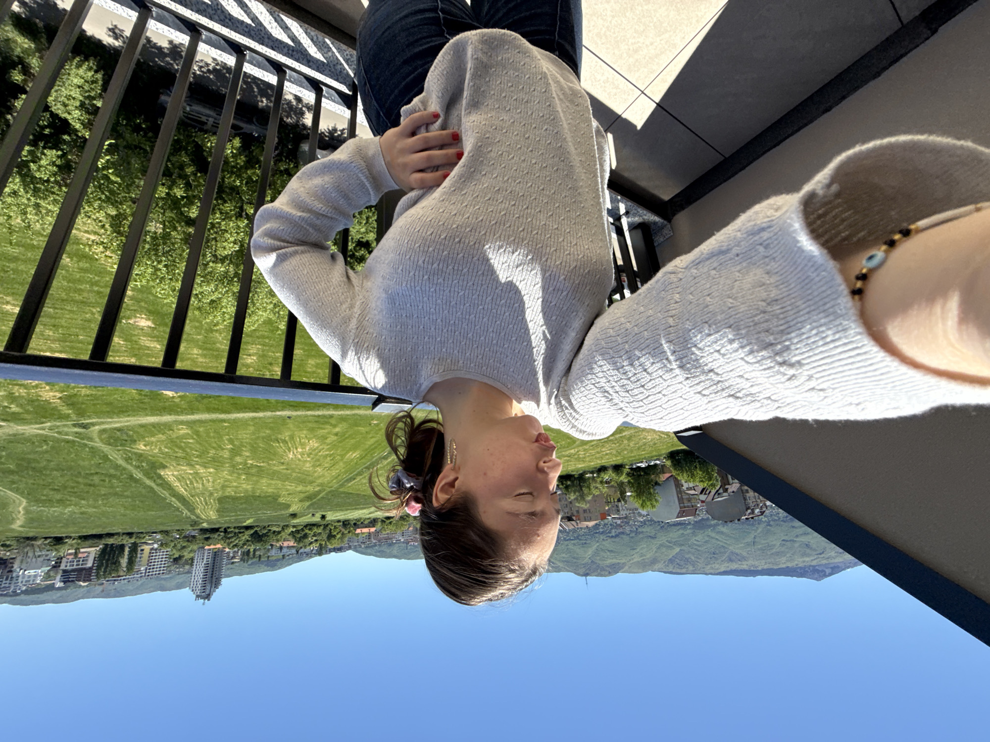 The image depicts a woman on a balcony overlooking a cityscape in Sofia, Bulgaria. She is wearing jeans and a white sweater and is positioned in the upper portion of the frame. Below her, a green field stretches towards buildings and mountains under a clear blue sky. A black balcony railing is visible on the left side of the frame. The woman is facing towards the ground with her eyes closed. A bracelet is visible on her wrist.