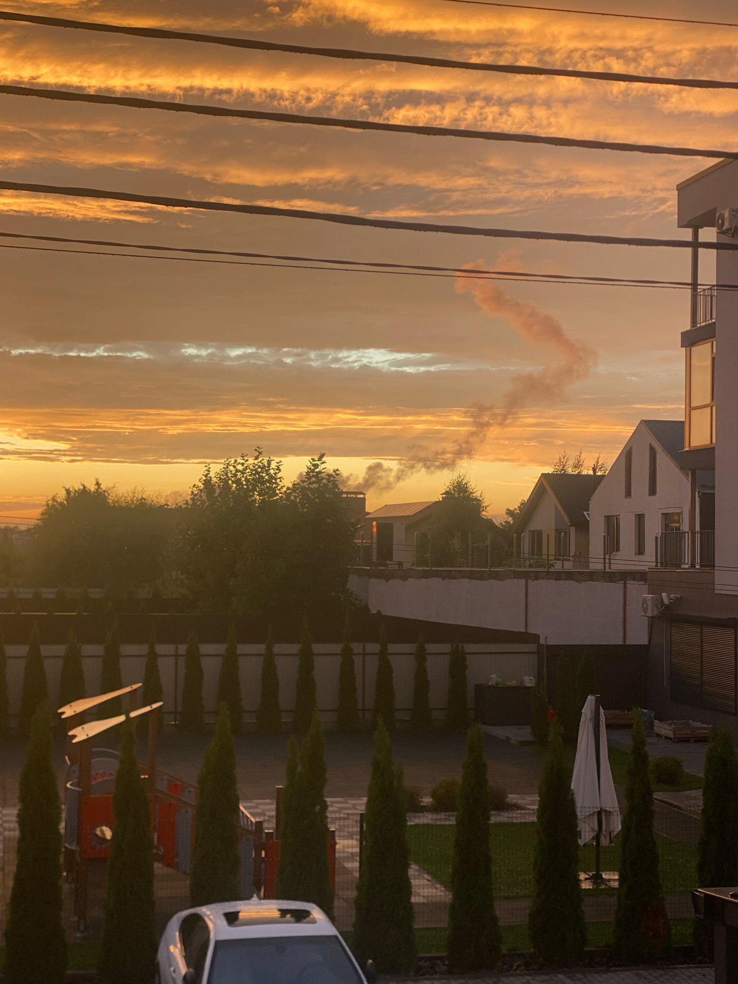 The image captures a serene sunset in Novi Petrivtsi, Ukraine. The sky is painted with hues of orange and yellow, with scattered clouds creating a dynamic texture. Several power lines stretch across the upper portion of the frame, interrupting the natural beauty of the sky. Below, a residential area is visible, featuring modern houses with clean lines and balconies. A plume of smoke rises from a chimney in the distance, suggesting human activity. In the foreground, a well-maintained yard with manicured cypress trees and a children's playground hints at a peaceful community. A white car with its sunroof open is parked in front of the trees, adding a touch of everyday life to the scene. The overall atmosphere is one of tranquility and domesticity, set against the backdrop of a vibrant sunset.