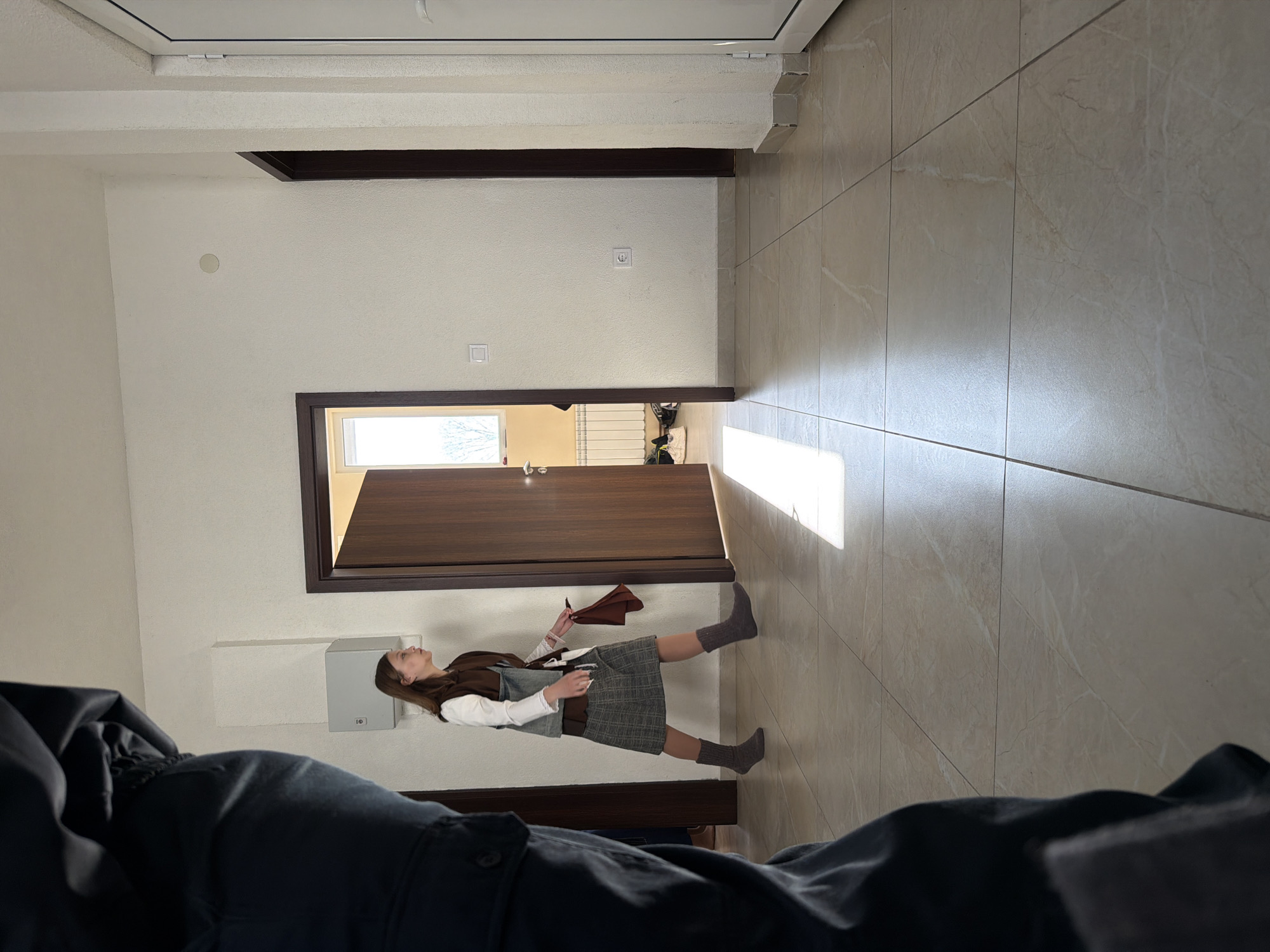 The image shows an indoor scene. A woman wearing a vest, dress, and socks stands near an open doorway in what appears to be a hallway. The walls are painted white, and the floor is covered in large beige tiles. A person, obscured and out of focus, is in the foreground wearing dark clothing. The location is potentially in Shumen, Bulgaria, although this is not directly verifiable from the image.