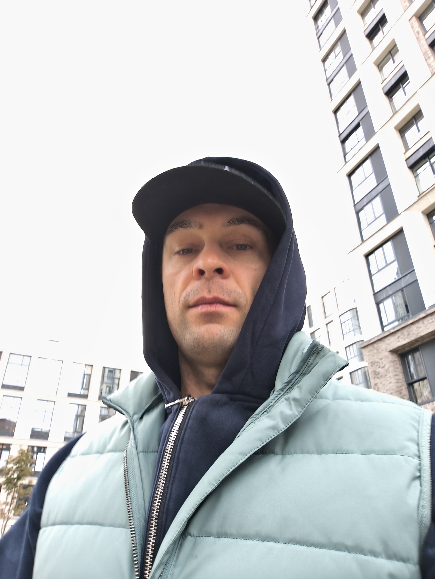 A man wearing a black cap, a navy hoodie, and a light green puffer vest takes a selfie outdoors. The man is centered in the frame. The background features apartment buildings, a cloudy sky, and some trees. Based on architectural style, the location is likely Moscow, Russia.
