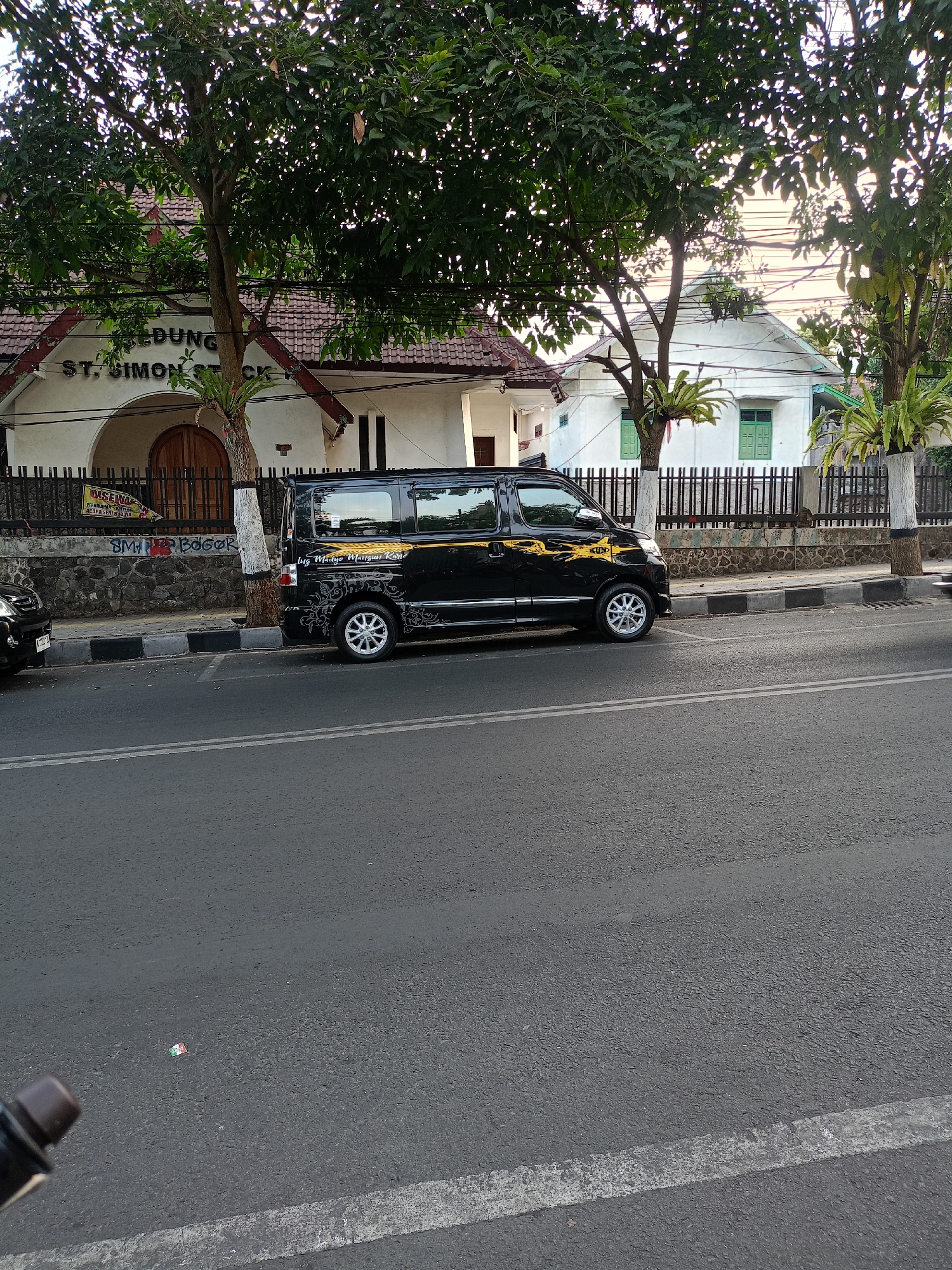 The image shows a street scene in the city of Batu, Indonesia. A black minivan is parked on the side of the road, with a building and trees in the background. The building appears to be a church or similar institution, with the words "GEDUNG ST. SIMON" visible on its facade. A fence separates the building from the street, and a sidewalk is present between the road and the fence. The sky is overcast, suggesting it might be morning or late afternoon. The street is paved, and there are visible lane markings. There are no people clearly visible in the image.
