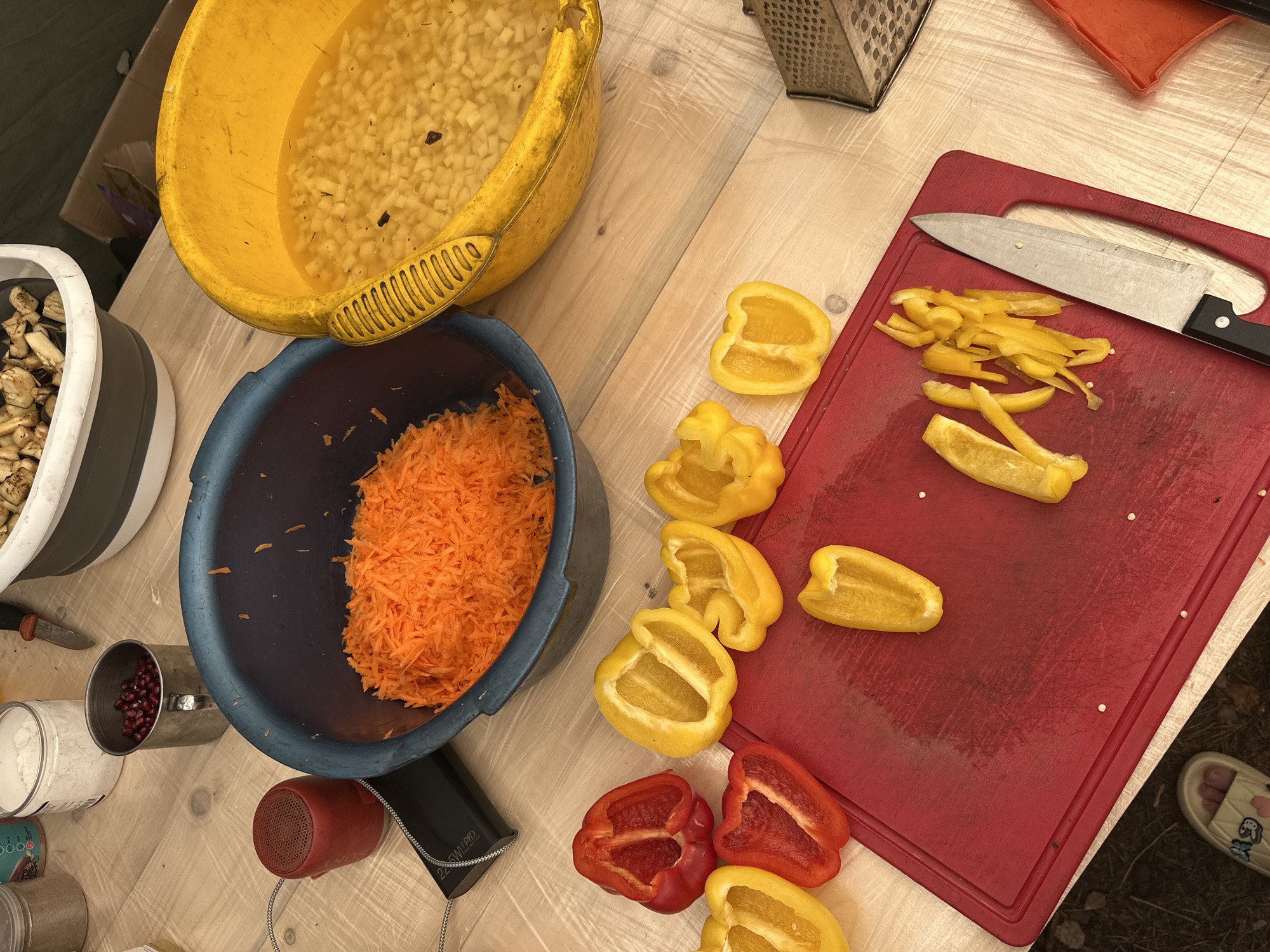 In a rustic setting on a wooden table, likely in Soanlakhti, Russia, a preparation for a meal is underway. The scene is dominated by colorful ingredients and cooking utensils. A large blue bowl contains a generous portion of shredded carrots. Adjacent to it, a bright yellow basin holds diced ingredients submerged in liquid.
In the foreground, a red cutting board is covered with sliced bell peppers. Several halves of red and yellow bell peppers are arranged on the table around the cutting board, some already cut into strips. A knife rests on the cutting board, positioned near the sliced peppers.
Other items visible include a small red speaker, a black portable charger, a metal cup filled with pomegranate seeds, and two jars containing white powders, possibly flour or spices. In the background, a grey and white collapsible basin holds chopped vegetables, likely eggplant. The overall impression is one of preparing a fresh, vibrant meal outdoors or in a simple kitchen.