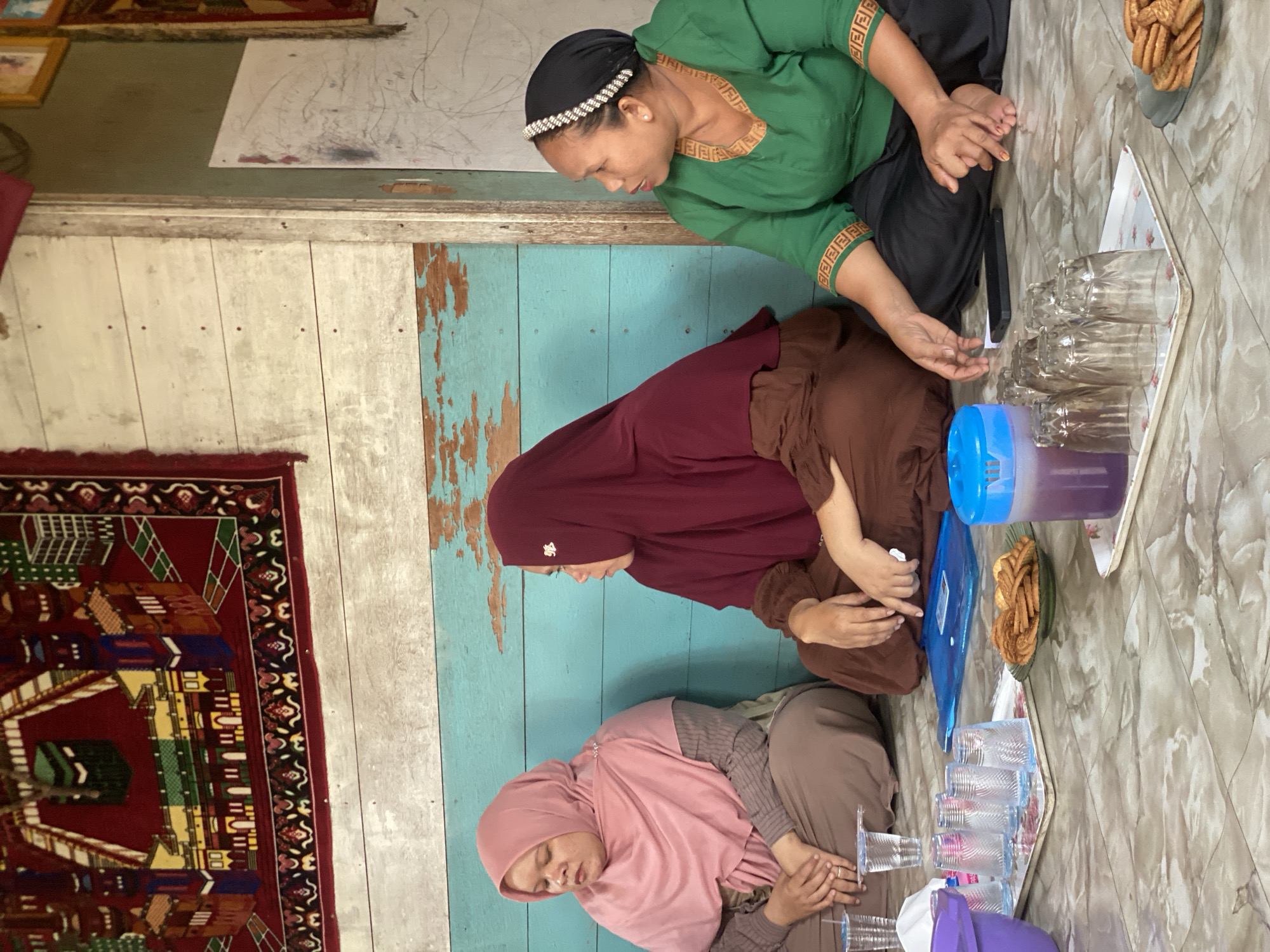 Three women are sitting on a floor in what appears to be a residential setting in Bawahan Selan, Indonesia. The woman on the left is wearing a pink hijab and brown clothing. The woman in the middle is wearing a maroon hijab and brown clothing. The woman on the right is wearing a black hijab with a jeweled headband and a green top. They are seated on a patterned floor, with a wall made of painted wooden planks behind them. A rug with a pattern depicting a mosque and buildings is hanging on the wall. In front of the women, there are trays with glasses, cookies, and a pitcher of purple liquid. The overall impression is that of a casual gathering or meeting.