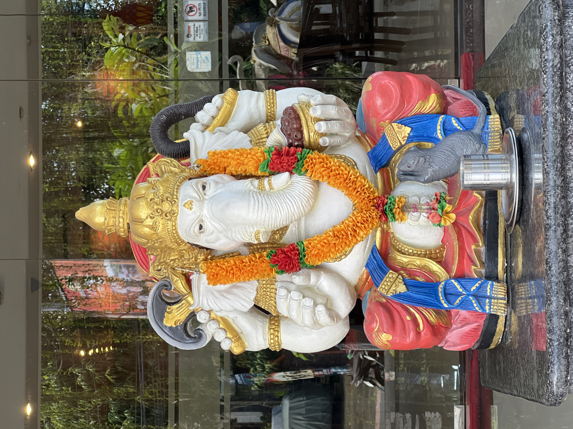The image shows a statue of the Hindu god Ganesha, with a garland of orange and red flowers around its neck. The statue is white with gold and blue accents, and it is sitting on a red base. A gray rat statue is also visible near Ganesha. The statue is located in the Ubud District, Indonesia. The statue is in front of a window, and reflections of trees and buildings are visible in the glass.