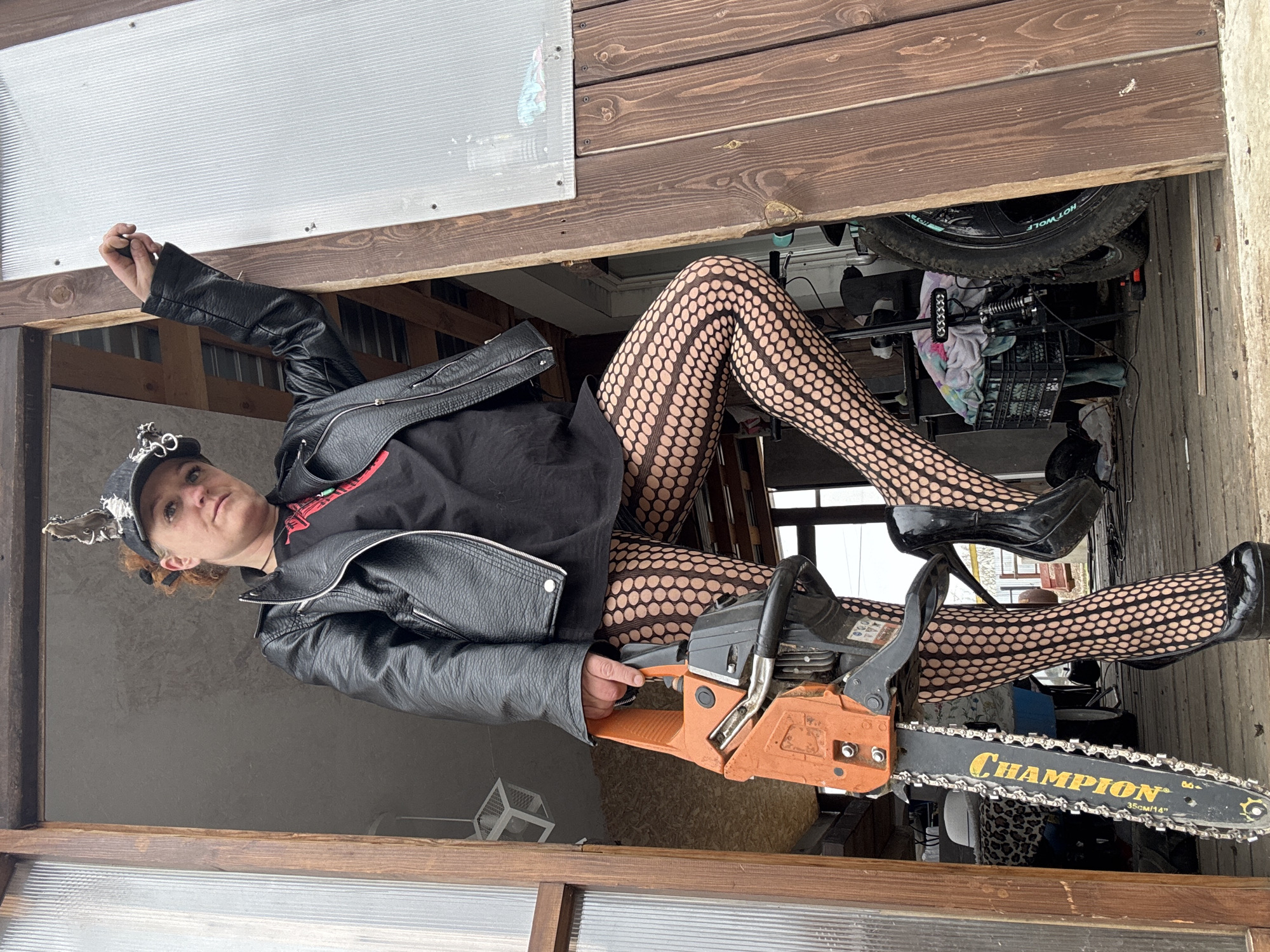 A woman is holding a chainsaw while sitting in a cluttered shed. She is wearing a black leather jacket, a black shirt, patterned fishnet stockings, and black high heels. She also has a black hat with metallic decorations on. The chainsaw is orange and black and has the word "CHAMPION" printed on the blade. The shed appears to be cluttered with various items, including a bicycle wheel, boxes, and other miscellaneous objects. The location is Chaikovsky, Russia.