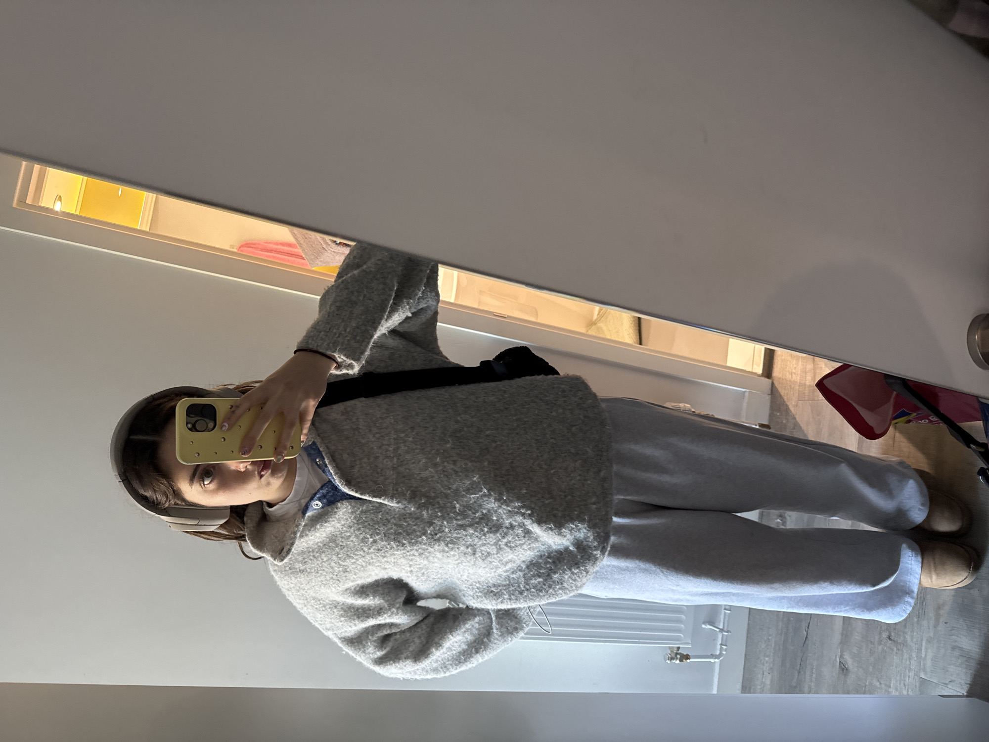 A mirror selfie captures a person, likely a young woman, standing indoors. The subject is wearing a light grey, fuzzy cardigan or jacket over a light-colored top with a visible collar, light grey loose-fitting trousers or sweatpants, and tan Ugg-style boots. Light-colored over-ear headphones are worn on her head. She holds a yellow smartphone with a studded case, obscuring most of her face except for her right eye and part of her mouth. Her fingernails are painted a light, possibly pink or white, color.
The background, reflected in the mirror, depicts a modern interior space. A white wall runs along the upper and left portions of the reflection. In the upper-middle, a horizontal rectangular recess or opening, possibly a shelf or cabinet, is brightly illuminated with a warm yellow light. Inside this recess, neatly folded pink and white items, appearing to be towels or clothing, are visible. To the right of the recess, a white wall with a silver door handle can be seen. The lower part of the reflection reveals light grey wooden or laminate flooring. A white radiator with exposed pipes is visible on the lower left side of the floor, and part of a red bag or basket is seen on the lower right, near the person's feet. The activity is a casual mirror selfie, likely taken in a home setting in Hull, United Kingdom. No visible text is present.
