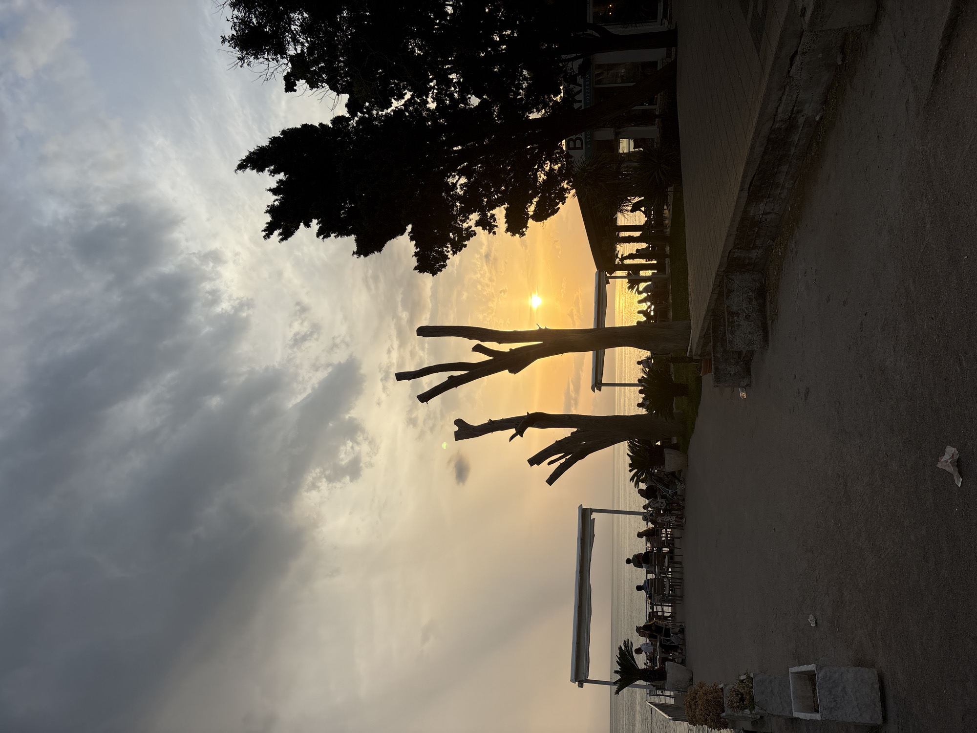 This image depicts a coastal scene in Bar, Montenegro, during sunset. The foreground shows a dark, paved walkway with planters and scattered debris. To the left, a seated group of people are visible at tables under a white canopy near the water's edge. Further to the right, a restaurant with a covered seating area is silhouetted against the setting sun. Tall, bare trees stand prominently in the midground, and dark silhouettes of trees line the right side of the frame. The sky is dramatic, with a mix of dark, heavy clouds and patches of lighter, hazy sky illuminated by the golden glow of the sunset. The sun is visible as a bright orb low on the horizon, casting reflections on the calm sea. Several individuals are seated at tables on the restaurant's patio, and some figures are walking. The overall atmosphere is serene, with the warm light of sunset creating a peaceful ambiance.