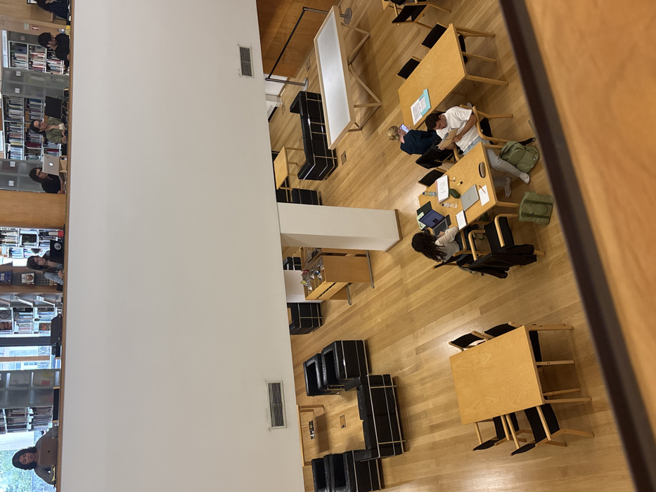 The image depicts a multi-level library interior within the Faculty of Arts and Humanities (Faculdade de Letras) building of the University of Porto, located in Lordelo do Ouro e Massarelos, Portugal.
The scene, viewed from an elevated perspective, features light-colored wooden flooring throughout. The upper level, delineated by a white wall and a wooden railing, shows a row of six individuals, predominantly women, seated at individual desks facing into the space. Each is engaged with a laptop computer. Behind them, bookshelves line the walls, visible through open sections.
The lower level is an expansive study and lounge area. In the foreground-midground, two large rectangular wooden tables with black-framed chairs are present. One table is empty with six chairs. The other table accommodates three individuals: a woman with dark curly hair using a laptop and tablet on the left, a man in a light shirt reading a book or document on the right, and a woman with blonde hair using a laptop in the background. Items on this table include laptops, notebooks, pens, a water bottle, and a green backpack.
Further into the lower level, two distinct lounge areas are arranged with black leather armchairs and sofas featuring chrome frames, each accompanied by a small wooden coffee table. A tall, multi-tiered wooden display unit containing various small objects stands near a central white support pillar. To the far right, a large rectangular table with a white top and a unique wooden base is visible. The far right wall is paneled with wood. White walls and ceilings complete the environment, with air vents visible on the white ledges. All individuals are focused on their respective tasks, indicating independent work or study.