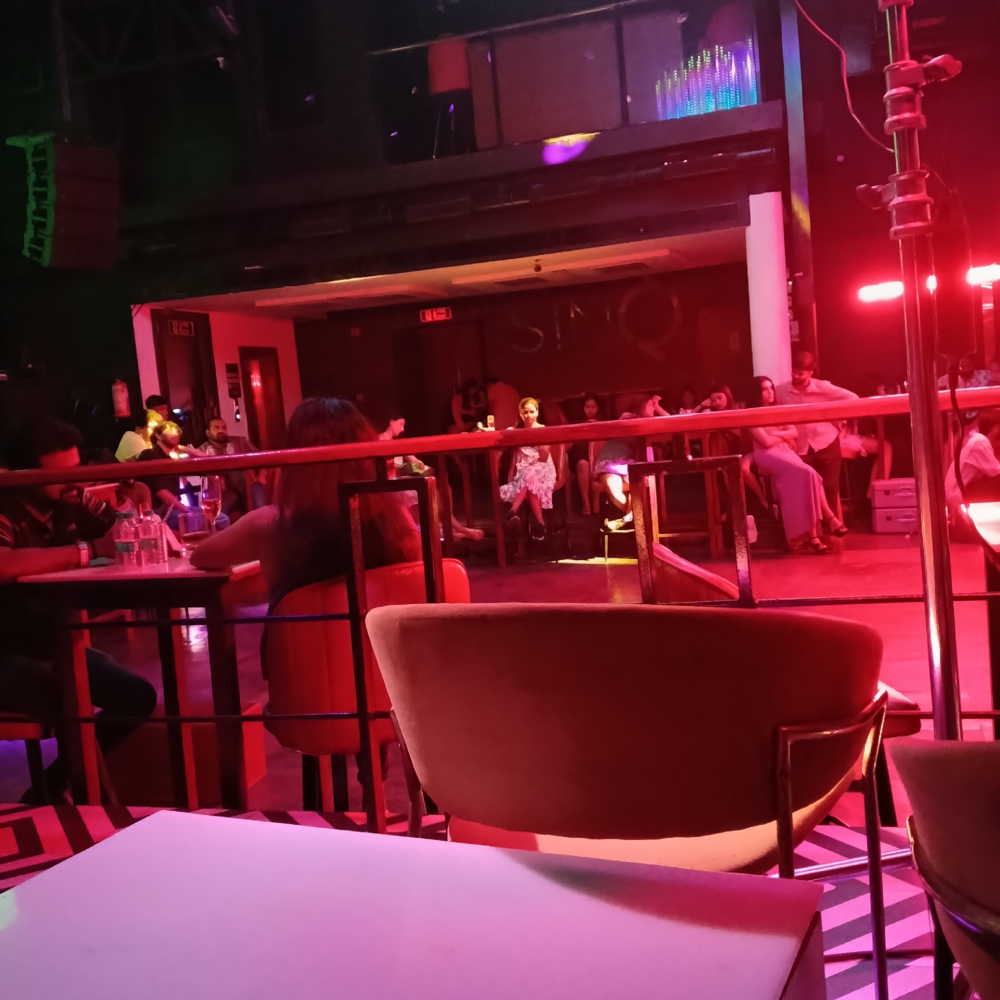 The image depicts the interior of what appears to be a nightclub or lounge in Sinquerim, India. The lighting is predominantly red and purple, creating a dim and atmospheric environment.
In the foreground, there are tables and chairs, some occupied. The chairs are upholstered and have metal frames. The tables are illuminated with a pinkish hue. The floor has a black and white striped pattern.
Behind the foreground, there is a low railing separating the seating area. Behind the railing, there are several people seated at tables or standing. The people are dressed in casual attire. Some are interacting with each other, while others appear to be observing the scene.
In the background, there is a partially visible sign that reads "SINO". Above the main area, there is a raised platform with additional seating and lighting. There are some light fixtures emitting a variety of colors. A speaker is visible in the upper-left.