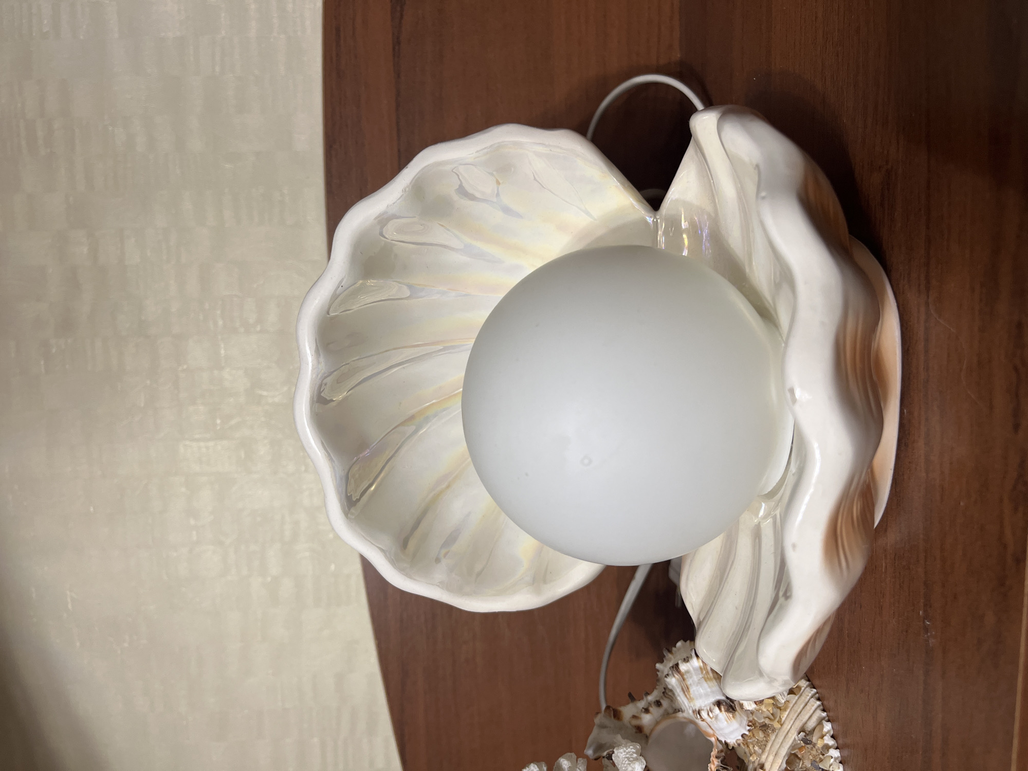 The image presents a close-up, vertically oriented shot of a decorative light fixture designed as an open clam shell. The main subject is a white, ceramic or porcelain shell sconce, showcasing a subtle iridescent, pearlescent finish with hints of pink, yellow, and blue on its inner surface. Nestled within the shell is a round, frosted white light bulb, which is unlit.
The background on the left features a light beige or off-white wall with a consistent, subtly textured, repeating pattern. To the right, a smooth, dark brown wooden surface is visible, likely part of furniture or wall paneling. White electrical wires extend from behind the shell fixture.
In the bottom right corner, a small collection of natural seashells is arranged. Some of these shells appear to be adorned with small granular materials or pebbles, adding to their texture. The scene depicts an indoor setting, suggesting a home environment with a coastal or beach-themed aesthetic. There are no people, activities, or visible text in the image, and the time of day is not discernible.