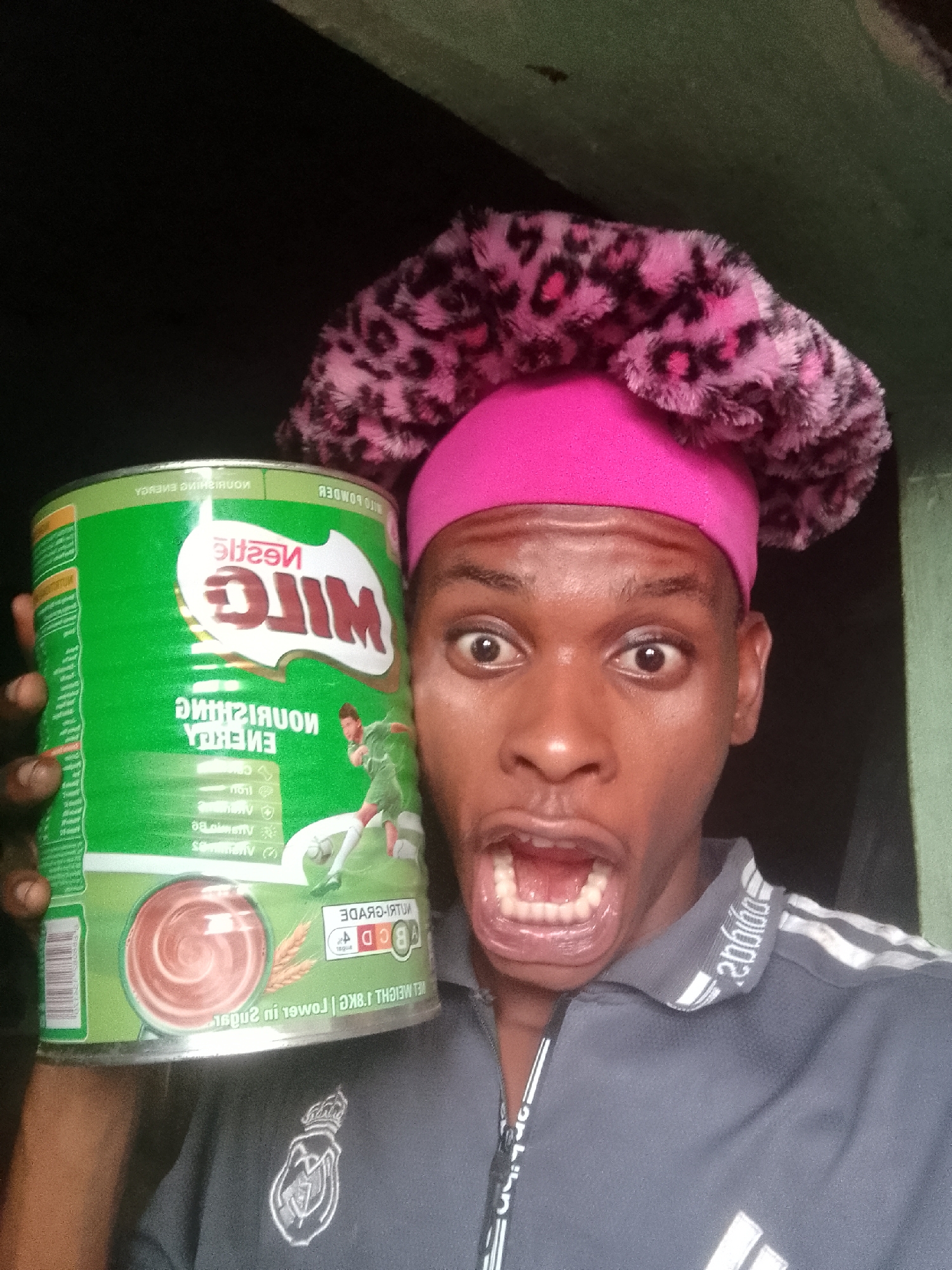 Here is a description of the image:
A close-up, selfie-style shot features a man holding a large green can of Nestle MILO. The can is predominantly green, with images of a soccer player and chocolate swirls, along with the brand's logo. The text on the can appears to be reflected, suggesting the image may be mirrored. The man has a surprised expression, with wide eyes and an open mouth. He's wearing a gray collared shirt with white Adidas stripes. On his head, he wears a pink and leopard print bouffant-style hat. The background is a mix of dark and slightly lighter green shades.