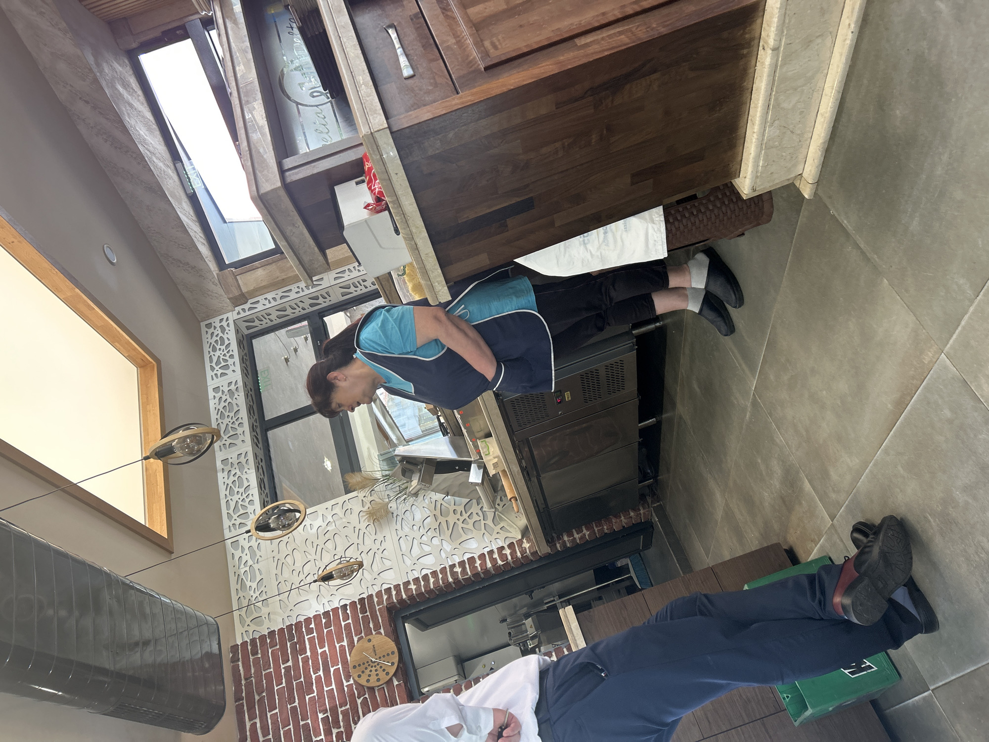 The image shows the interior of a commercial space, possibly a restaurant or bakery, located in Kazanlak, Bulgaria. Two individuals are present. One is an employee wearing a light blue shirt and a dark blue apron. The other individual is wearing a white shirt and dark blue pants. The space features a combination of architectural elements, including brick walls, decorative lattice panels, a window with a view to the outside, and wooden cabinets. The floor is tiled. Lighting fixtures are suspended from the ceiling. Commercial kitchen equipment is visible, including a refrigerator unit.