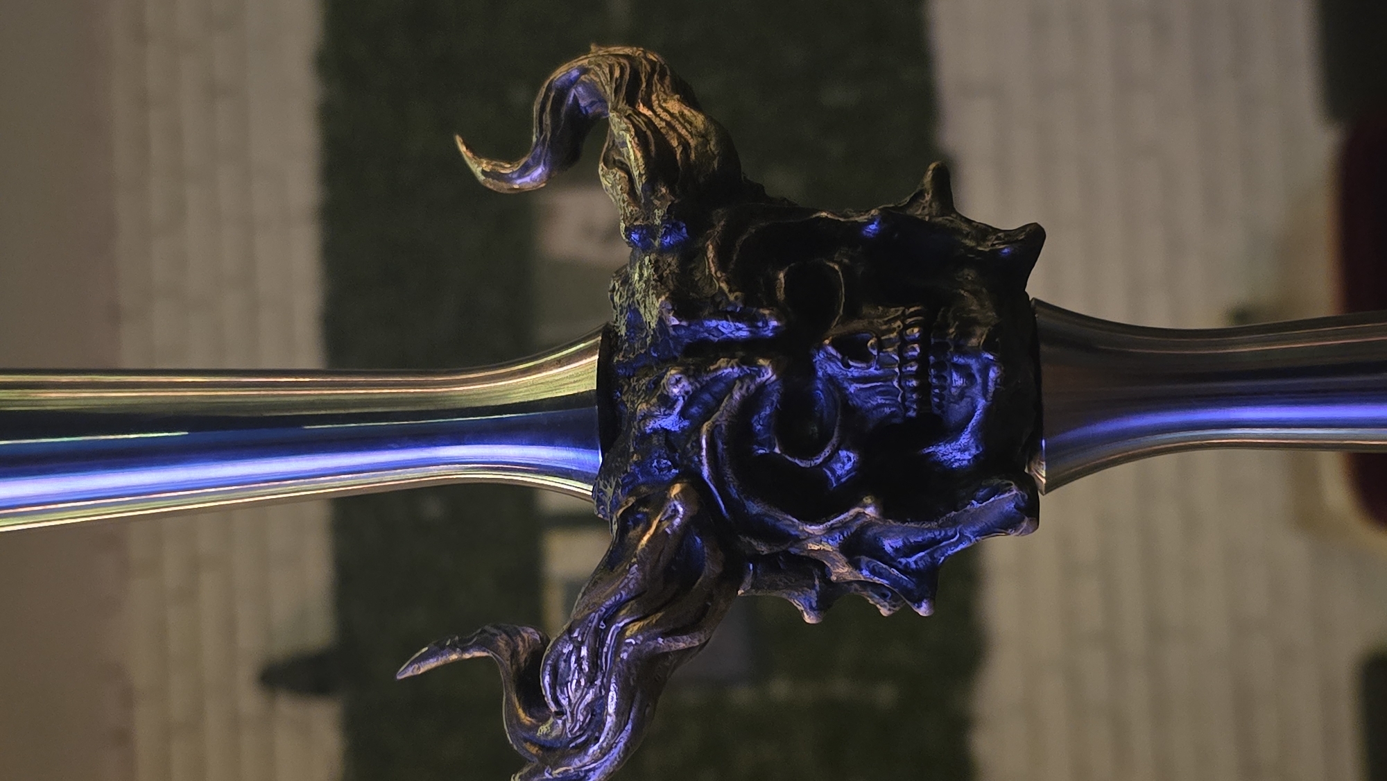 The image displays a close-up, horizontal view of an ornate, dark metallic object, strongly resembling the hilt of a sword or a ceremonial weapon. The central feature is a highly detailed, sculpted guard or pommel designed like a monstrous or demonic skull, complete with intricate, swirling organic forms or horns extending upwards and downwards. The material appears to be dark metal with textured surfaces, highlighted with accents of bronze or gold.
Two sleek, cylindrical elements extend horizontally from the central skull-like hilt, appearing to be blades or shafts. These elements are highly reflective and smooth, intensely catching and refracting a vibrant blue light. This blue illumination bathes parts of the skull sculpture as well, creating strong contrasts against the shadowed recesses and the warmer bronze accents. The dramatic lighting lends an eerie and mystical atmosphere to the object.
The background is intentionally blurred and out of focus, providing a soft, indistinct backdrop. On the left, a lighter, beige or pale grey area with a subtle vertical pattern is visible, while the right side shows a darker, greenish, ambiguous shape. No activity is taking place; the image is a static portrayal of this meticulously crafted object. There is no visible text in the image. The overall aesthetic is dark fantasy or gothic.