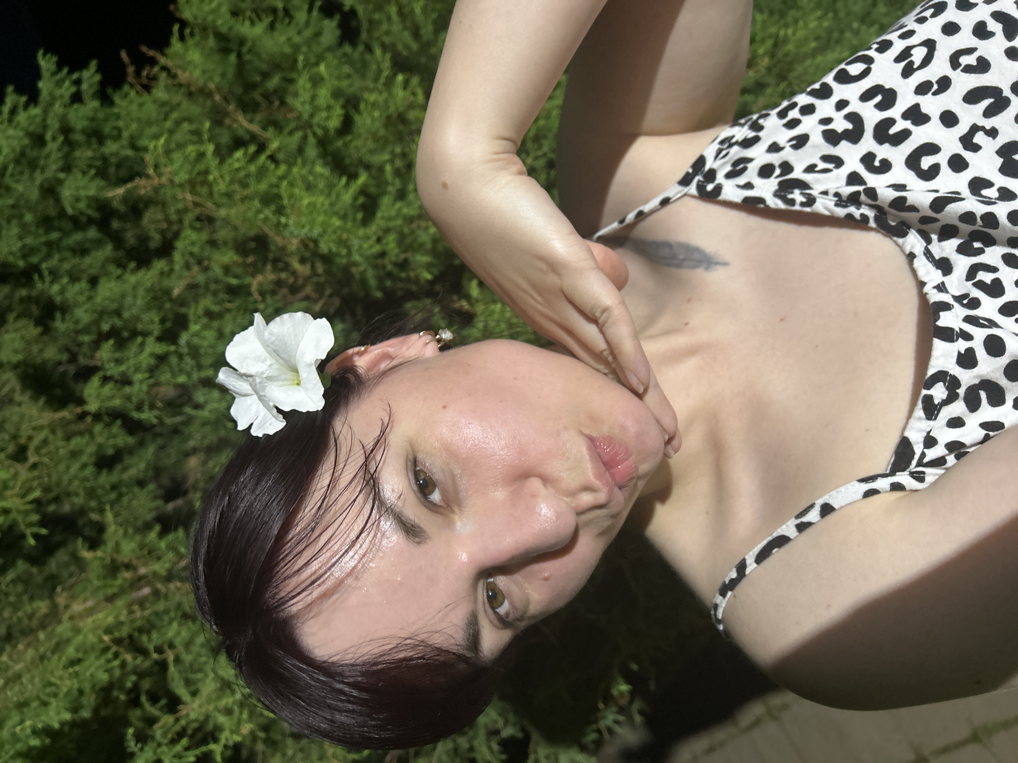A young woman with dark hair adorned with a white flower poses for a selfie outdoors. She is wearing a white and black leopard print camisole and has her hand gently placed on her neck. The background consists of lush green foliage, possibly from a tree or shrub, and a dark, out-of-focus area that suggests it is nighttime or a dimly lit environment. There is a faint tattoo of a feather visible on her upper chest. The lighting in the image casts a shadow across her left shoulder and arm. The location in the city of Фоки, Russia is not explicitly identifiable from the visual information provided.
