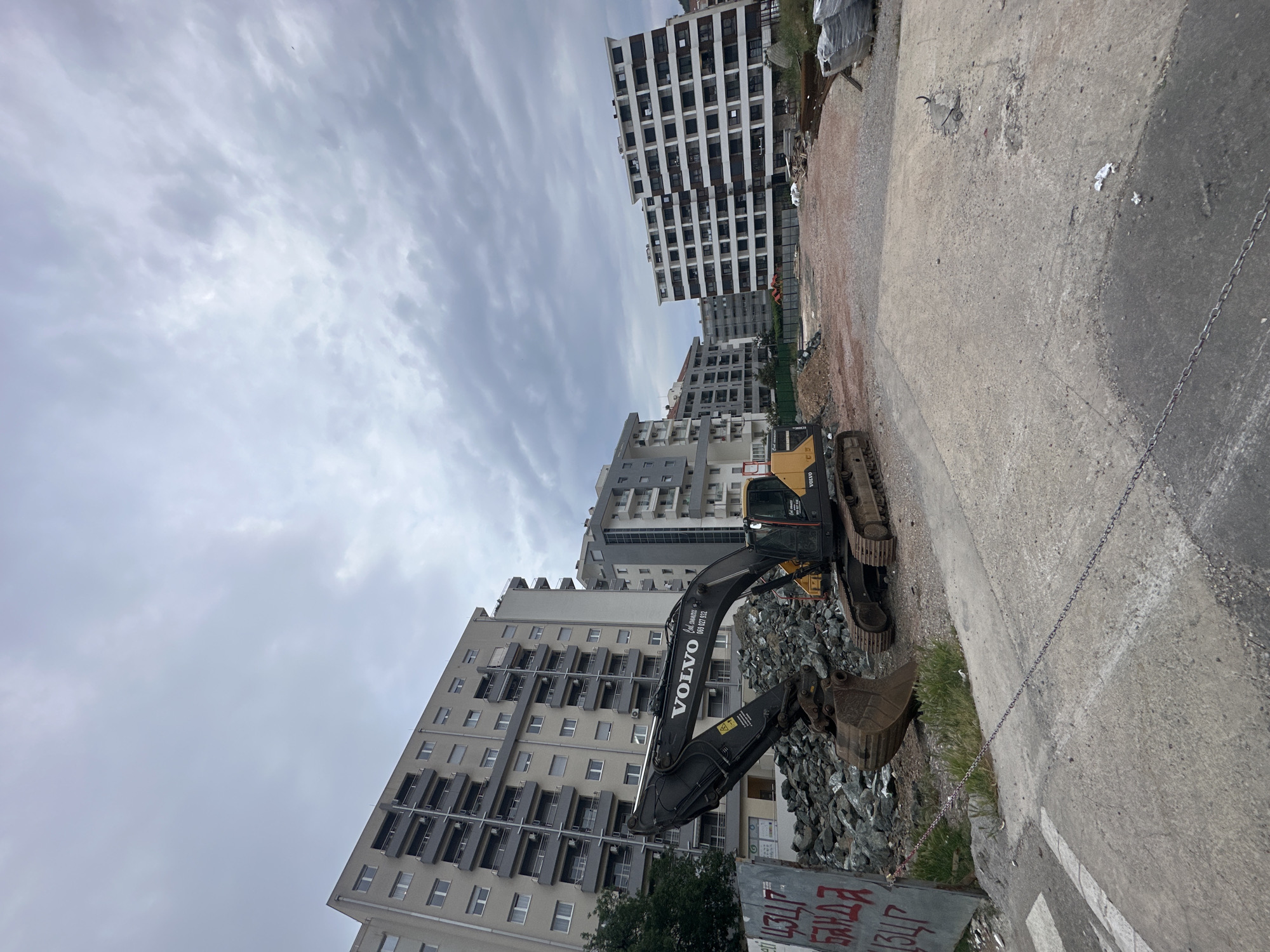 An excavator is parked on a construction site in Budva, Montenegro. The machine is angled towards a pile of rocks and rubble. Several tall, modern apartment buildings stand in the background, reaching up into a cloudy sky. A chain runs along the edge of a concrete surface, delineating the boundary of the construction zone.