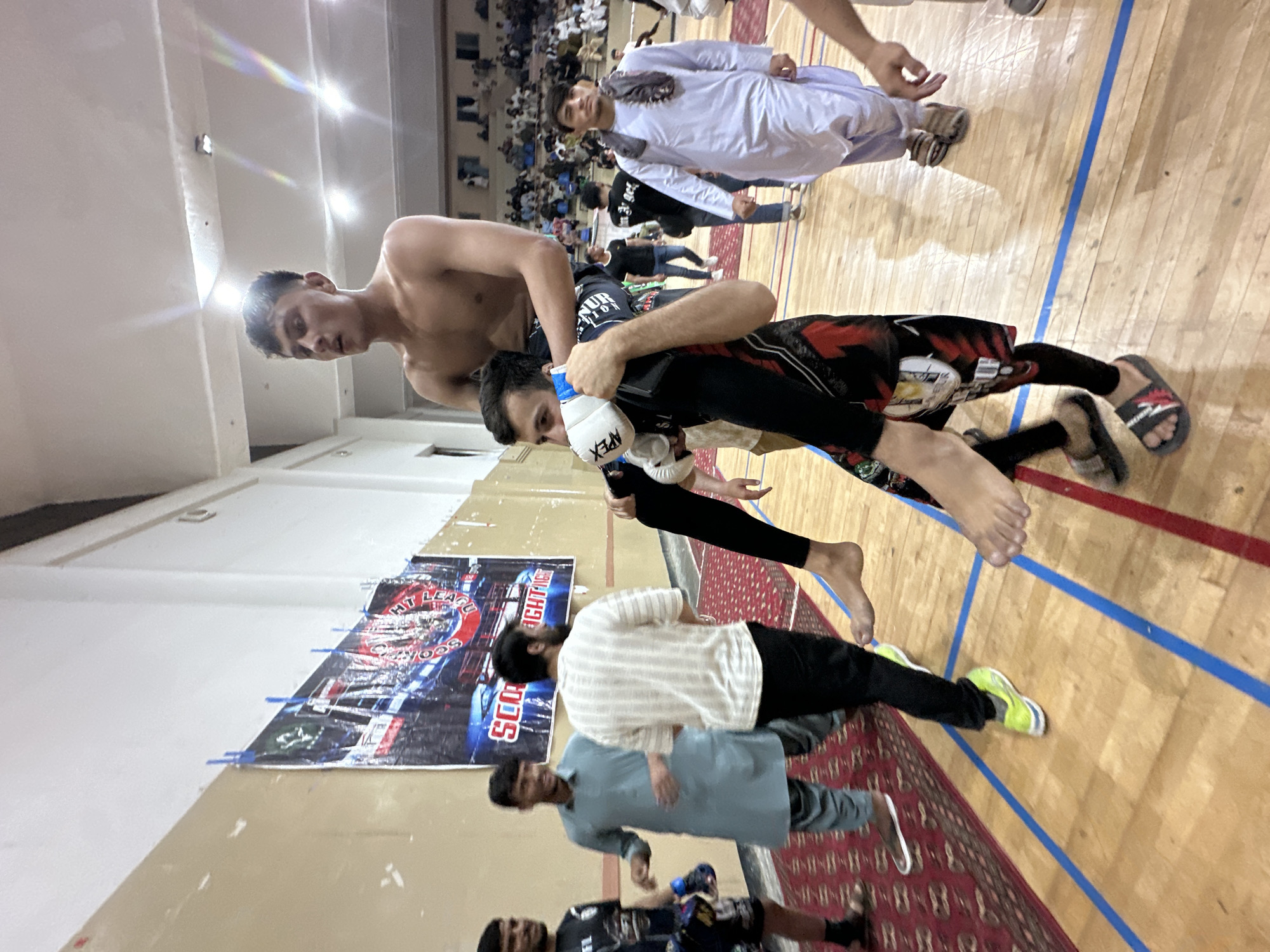 The image shows two men engaged in a grappling martial arts competition on a wooden floor marked with blue lines. One man is shirtless and has dark hair, while the other is wearing MMA gloves and clothing. They are near a banner that reads "SCOUTING FIGHT LEAGUE." A crowd is visible in the background. The location is Quetta City Tehsil, Pakistan.