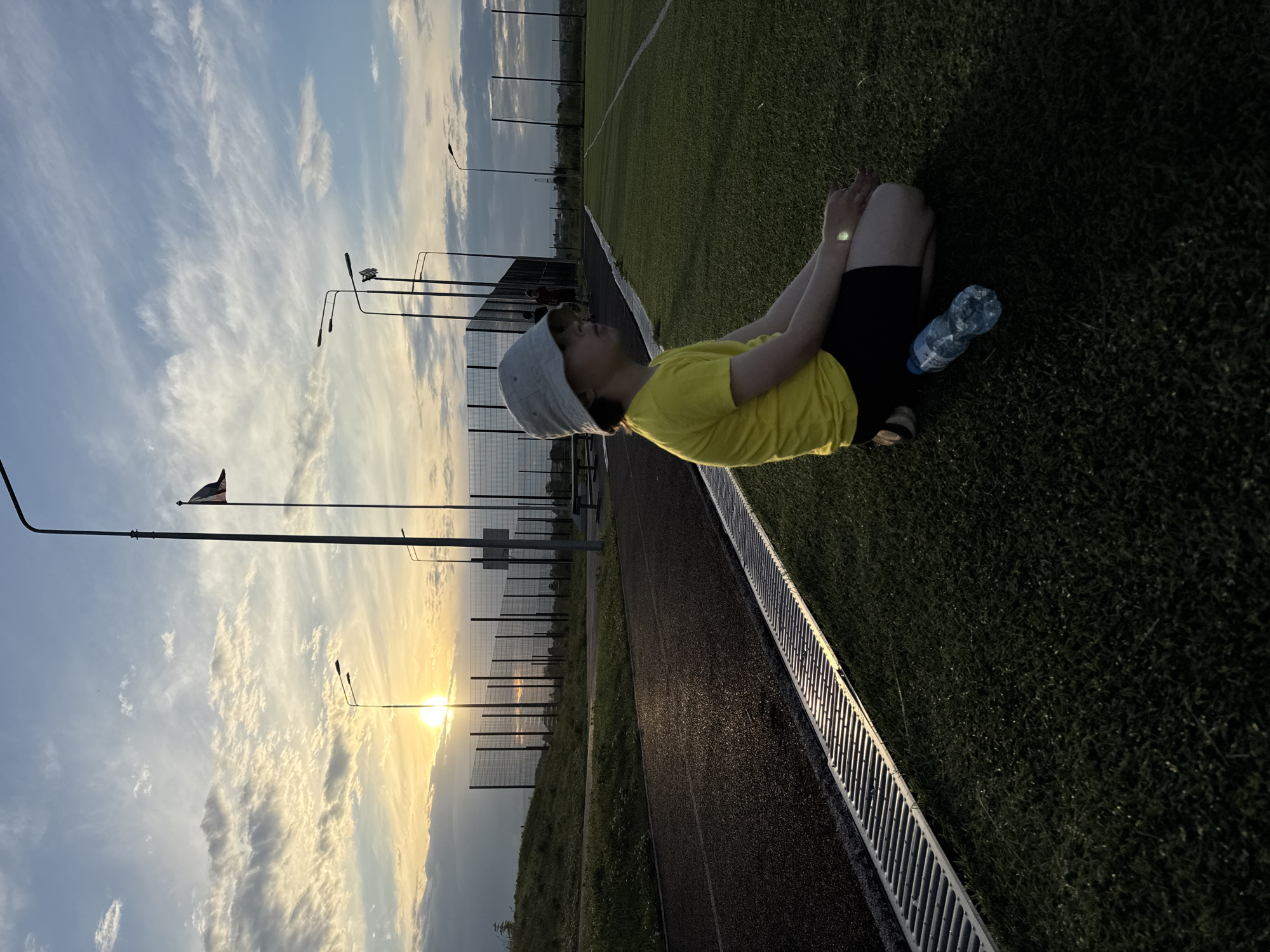 The image shows a young person in a yellow t-shirt and black shorts sitting on green grass near a running track. They are wearing a grey bucket hat and have their legs bent with their hands clasped together. A plastic water bottle is placed on the grass next to them. The background features a soccer field with white lines, a fence, and several lampposts. The sky is blue with wispy clouds, and the sun is setting, casting a warm glow over the scene. The overall impression is one of relaxation or a break during physical activity. The location appears to be an outdoor sports facility in Gornoe, Russia, given the context.