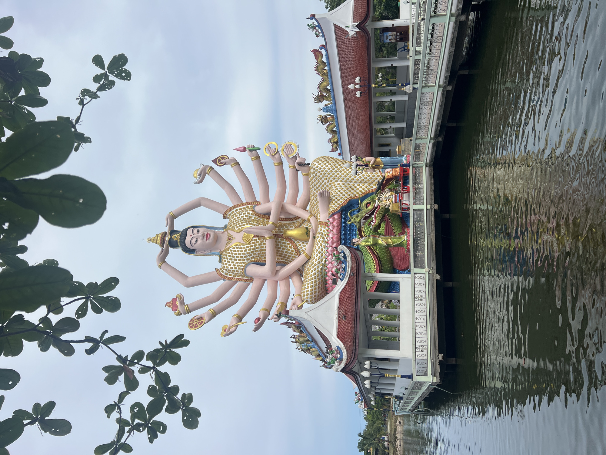 A large, multi-armed statue of a seated deity, likely a Buddhist figure, dominates the center of the image. The statue is adorned in a gold and white patterned robe and wears an elaborate crown. It is positioned on a raised platform that extends into a body of water. This platform supports a building with a red-tiled roof, decorated with ornate dragon figures and traditional architectural elements. The building has white walls and open colonnades. A walkway with a green railing connects parts of the platform. In the water, the reflection of the statue and the building is visible, with ripples distorting the image. Trees with green leaves hang into the frame from the top, partially obscuring the sky. The sky is overcast with a light gray hue. The scene is located at Baan Plai Laem, Thailand.