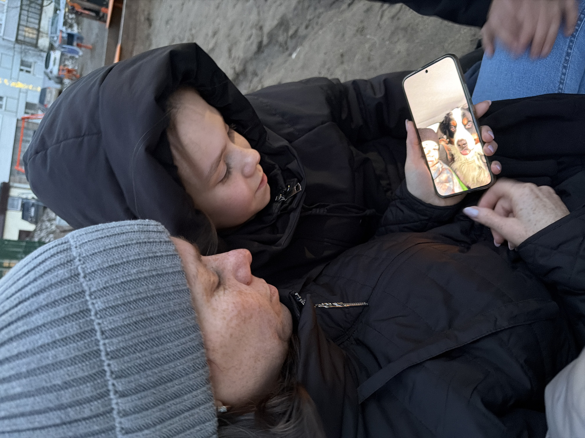 Two individuals are reclining or lying down outdoors, viewed from a slightly elevated, tilted perspective. In the foreground, an adult person, likely a woman, is wearing a grey ribbed knit beanie and a black puffer jacket. Her face, featuring freckles, is turned upwards, looking towards the person beside her.
Beside her, a younger individual, possibly a child or young teen, is also wearing a black hooded puffer jacket, with the hood up. This person is holding a black smartphone in their right hand, showing the screen to the adult. The phone's screen displays a close-up image of a large dog, possibly a Bernese Mountain Dog, with its tongue slightly out, and partially visible next to it, the head of a person (potentially a child). Both main subjects appear to be focused on the phone's content.
Partially visible in the background, to the right, are the hands and denim-clad legs of a third person, suggesting companionship. The outdoor setting features a ground of sand or dirt. In the distant background, urban structures including buildings with multiple windows and what appears to be a playground or construction equipment are visible under a bright, possibly overcast, sky. The warm clothing suggests cool or cold weather. The scene conveys a moment of shared attention and intimacy in an outdoor environment in Chaikovsky, Russia. There is no legible text visible in the image.