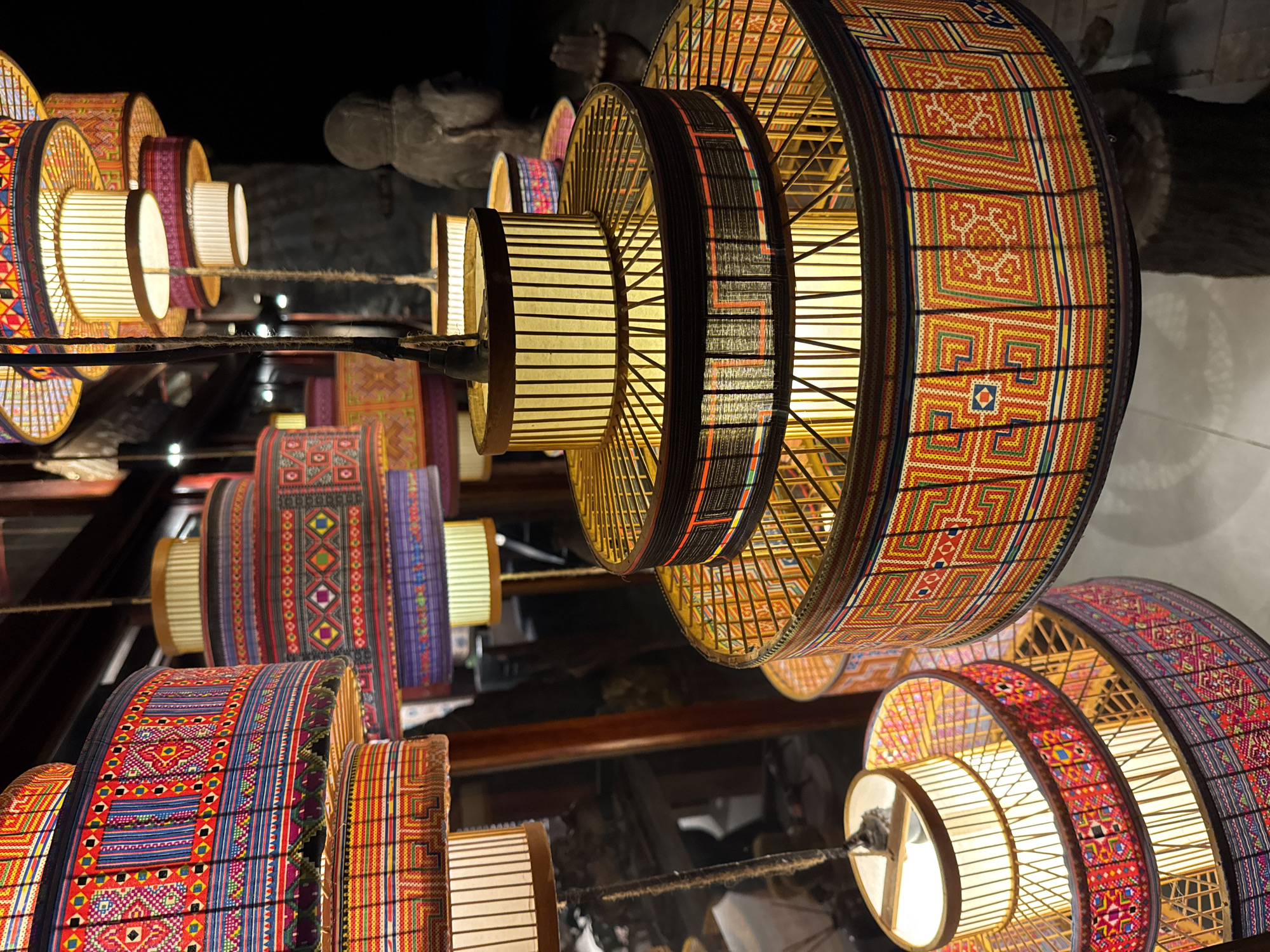 This image depicts a collection of traditional, handcrafted lamps, likely made of bamboo and fabric, displayed in an indoor setting in Moc Chau, Vietnam. The lamps feature intricate, colorful geometric patterns in shades of red, blue, yellow, and purple. They vary in size and shape, with some appearing cylindrical and others tiered, resembling pagodas or stacked bowls. Several lamps are illuminated from within, casting a warm glow through their translucent panels. In the background, a stone statue of a figure with a hood and clasped hands is visible, along with other dimly lit decorative elements. The overall ambiance suggests a retail or exhibition space, possibly a shop selling local crafts. The presence of what appears to be a furry animal at the bottom right corner adds an unexpected element to the scene. The lighting is dim, with the lamps being the primary light sources, creating a cozy and atmospheric environment.
