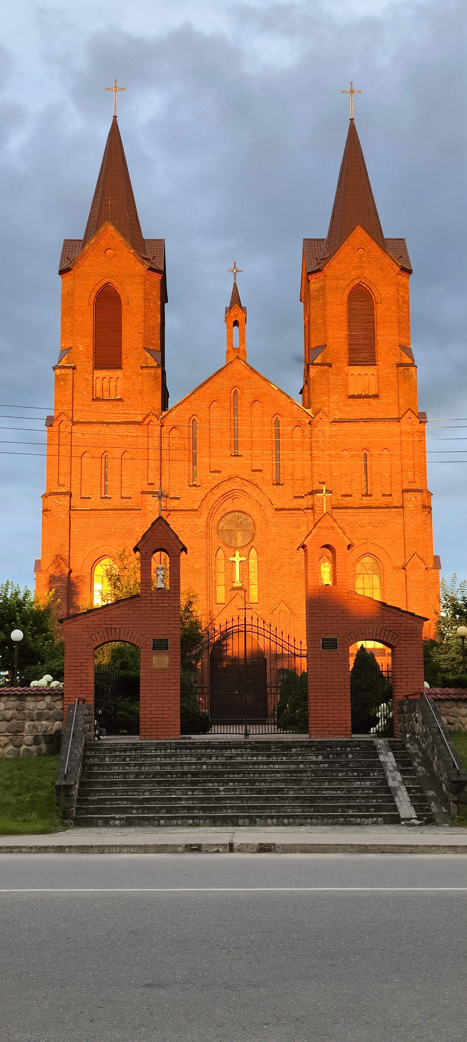 A low-angle shot of the Church of the Assumption of the Virgin Mary in Myory, Belarus, bathed in warm sunset lighting. The church is constructed of red brick with two prominent towers and a central facade adorned with a crucifix. In front of the church, a brick gateway with wrought-iron gates leads to a stepped entrance. The foreground consists of a road. The sky is overcast, with grey clouds contrasting the warm hues of the church.