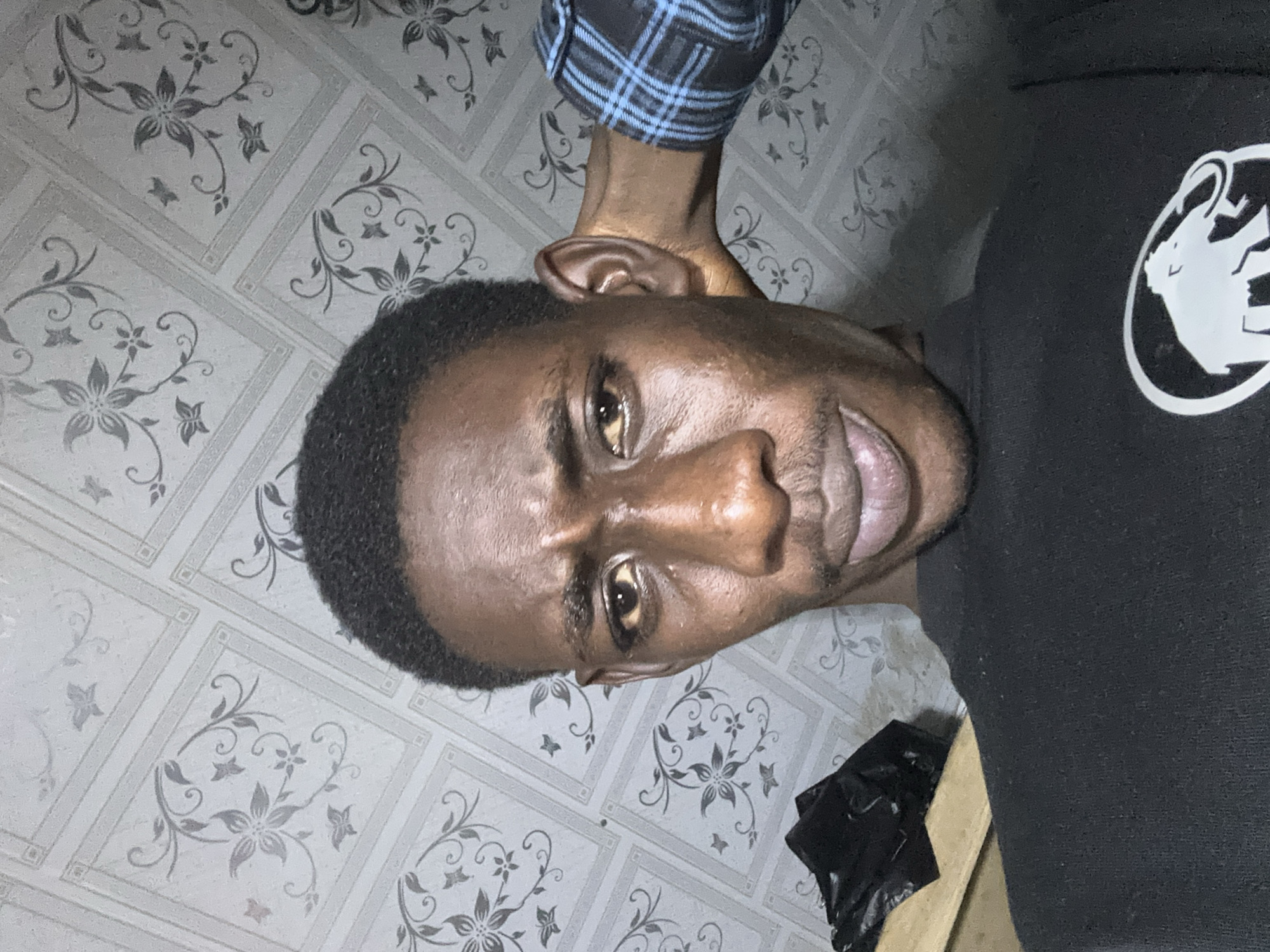 A young Black man is looking at the camera with a strained expression. He has dark skin and short, black hair. He is wearing a black t-shirt with a white logo of an elephant in a circle on the left side of his chest. His right arm is bent, with his hand behind his head. He is wearing a blue and black plaid shirt on his right arm. The background is a wall covered with patterned wallpaper. The wallpaper has a white background with a repeating pattern of grey floral designs and butterflies. To the left of the man, a black plastic bag is visible on a wooden surface. The image appears to be taken indoors. There is no identifiable location in the city of Durumi, Nigeria.