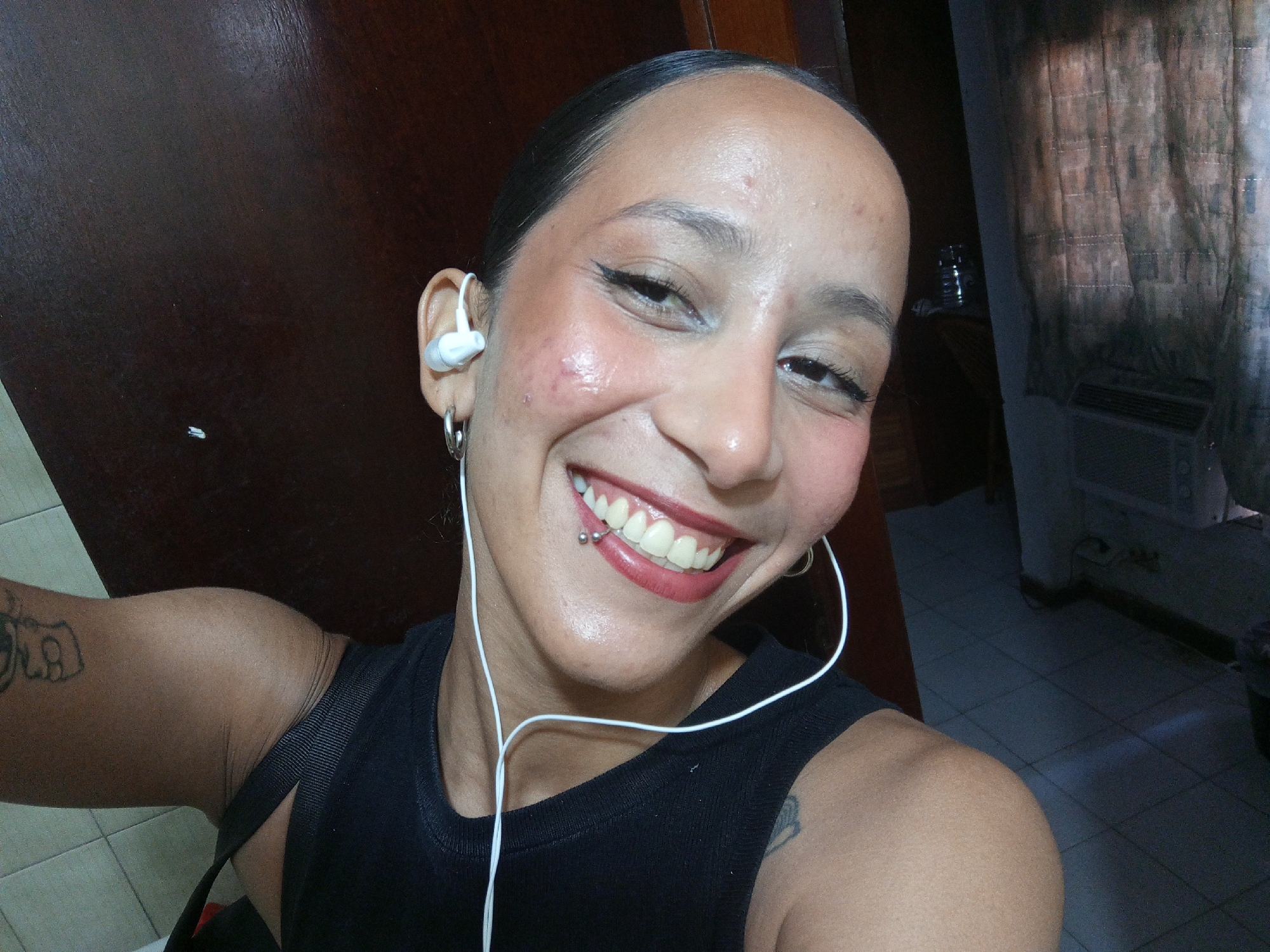 A medium shot captures a person with dark, slicked-back hair, fair skin, and a bright smile revealing a lip piercing on the lower lip. They are wearing a black tank top and white earphones. Tattoos are visible on their upper arms. The background includes a dark wooden door on the left and a patterned curtain near an air conditioning unit on the right. The floor appears to be tiled. The specific location within Porlamar, Venezuela, cannot be determined from the visual information provided.