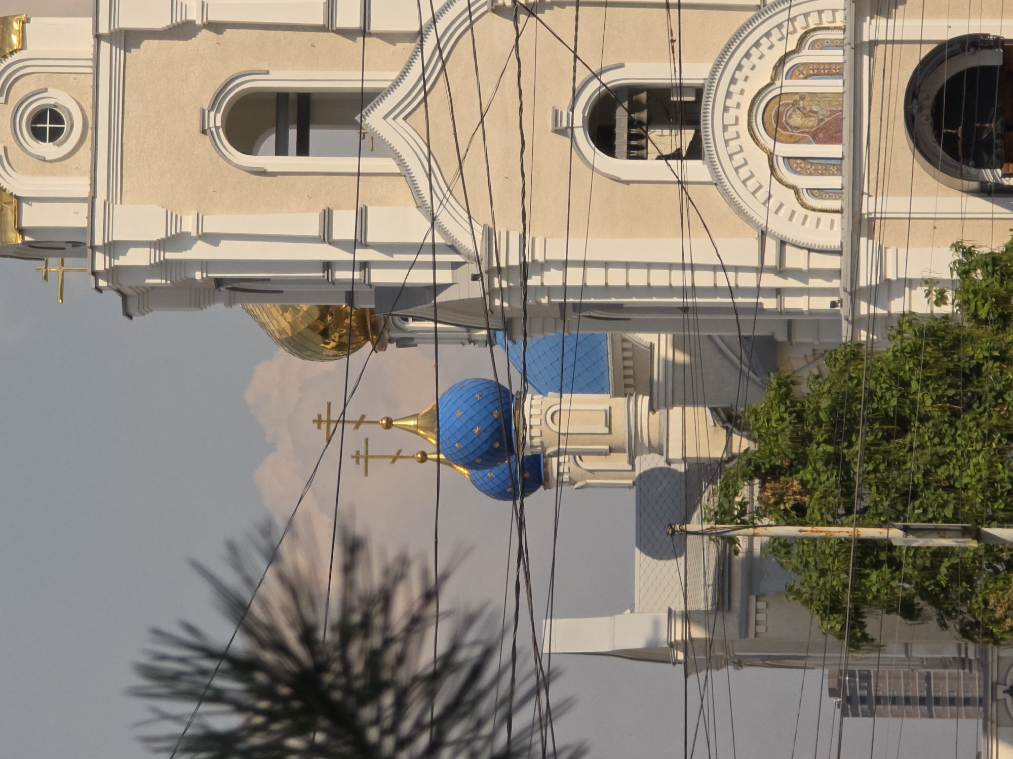 A segment of a cream-colored church with white trim and blue domes is visible on a sunny day, partially obscured by tree branches and numerous electrical wires. The church features golden crosses atop its domes and a circular window on its upper facade. Below the window, an arched opening contains bells. Further down, an arched mosaic depicts the Virgin Mary holding the infant Jesus. In the background, a modern brick building rises, and the sky is a pale blue with scattered clouds. The image was captured in Rostov-on-Don, Russia.