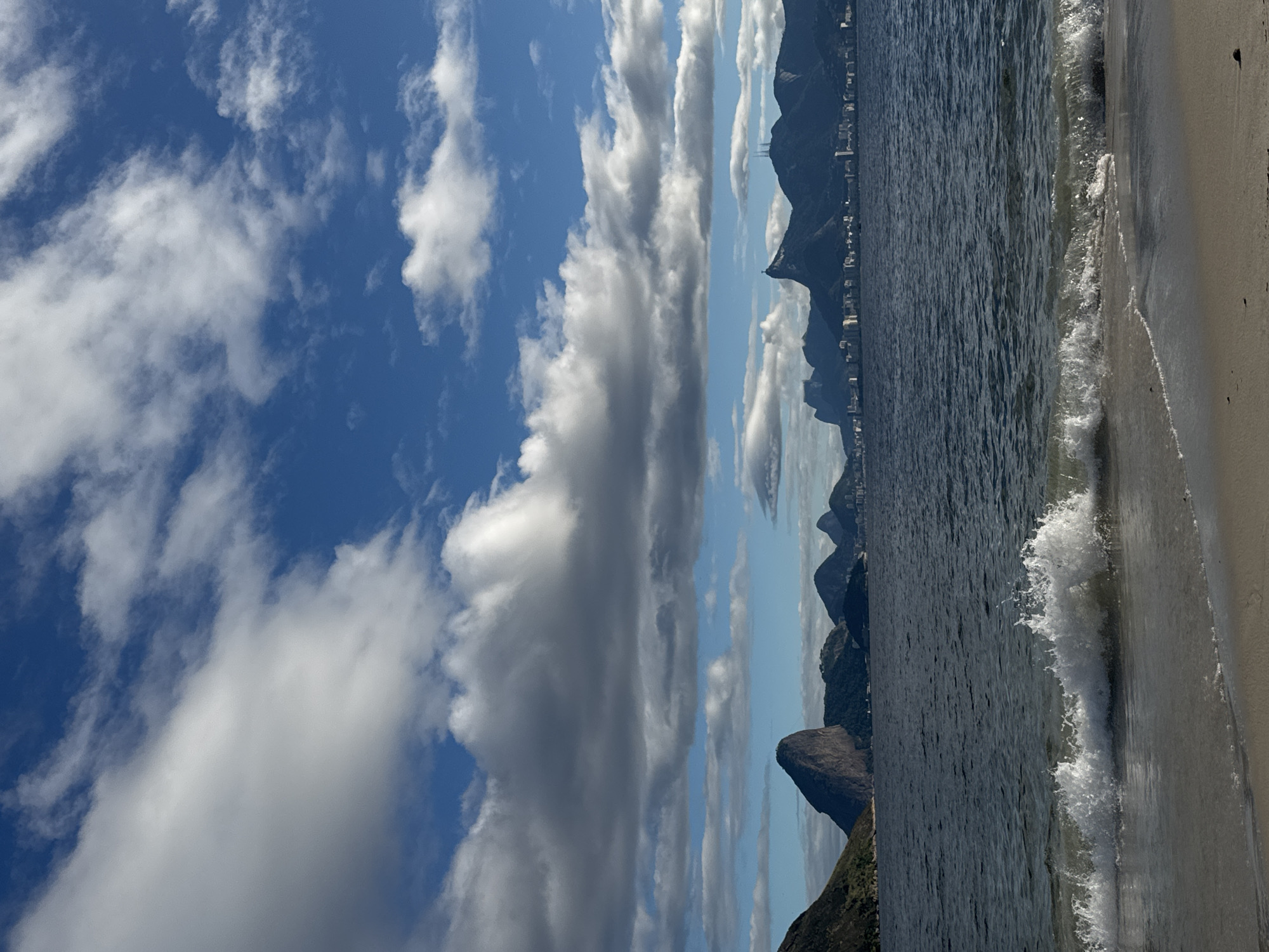 This is a vertical, eye-level, outdoor, medium shot photograph taken at a beach in Niterói, Brazil. The image depicts a coastal landscape with a wide expanse of deep blue water, a sandy beach in the foreground, and a mountainous coastline in the background. The sky is a vibrant blue, filled with a mix of large, fluffy white clouds and darker, more scattered grey clouds. Waves are gently rolling onto the shore, creating white foam and a sense of calm.
The distant coastline features a series of dark, lush green mountains. On one of the mountains, the iconic statue of Christ the Redeemer is visible, serving as a clear location cue for Rio de Janeiro and its surrounding areas, which include Niterói. Buildings are scattered along the base of the mountains, suggesting a populated urban area adjacent to the natural landscape.
The lighting suggests it is daytime, likely late morning or early afternoon, given the bright sky and the way the light reflects on the water. The weather appears pleasant, with a mix of sun and clouds. No people or specific objects are the primary subjects of the image, focusing instead on the natural beauty of the scene and its iconic landmarks. There is no visible text in the image. The overall mood conveyed is one of tranquility and natural grandeur.