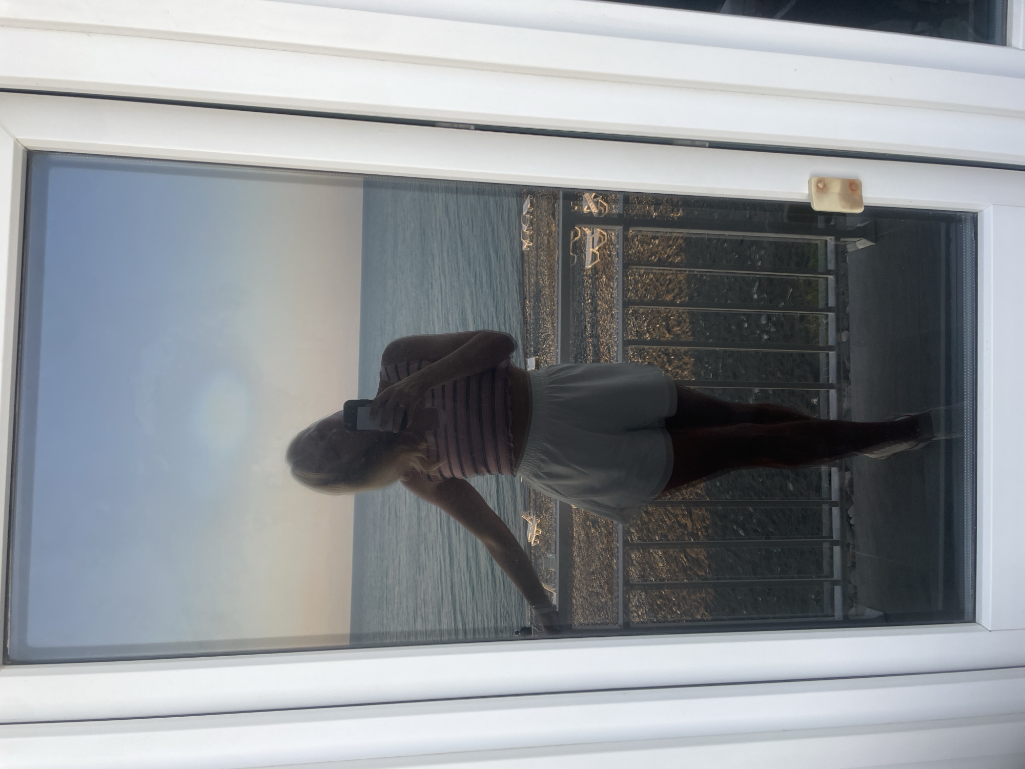 The image shows a reflection in a glass door of a woman taking a selfie. She is standing on a balcony overlooking a beach and the sea. The sky is clear with the sun setting. The woman is wearing a striped crop top and white shorts. On the beach, there are several empty lounge chairs. The scene is captured from a slightly elevated perspective, suggesting the balcony is on an upper floor. The location is identified as Giachrypsh, Abkhazia.