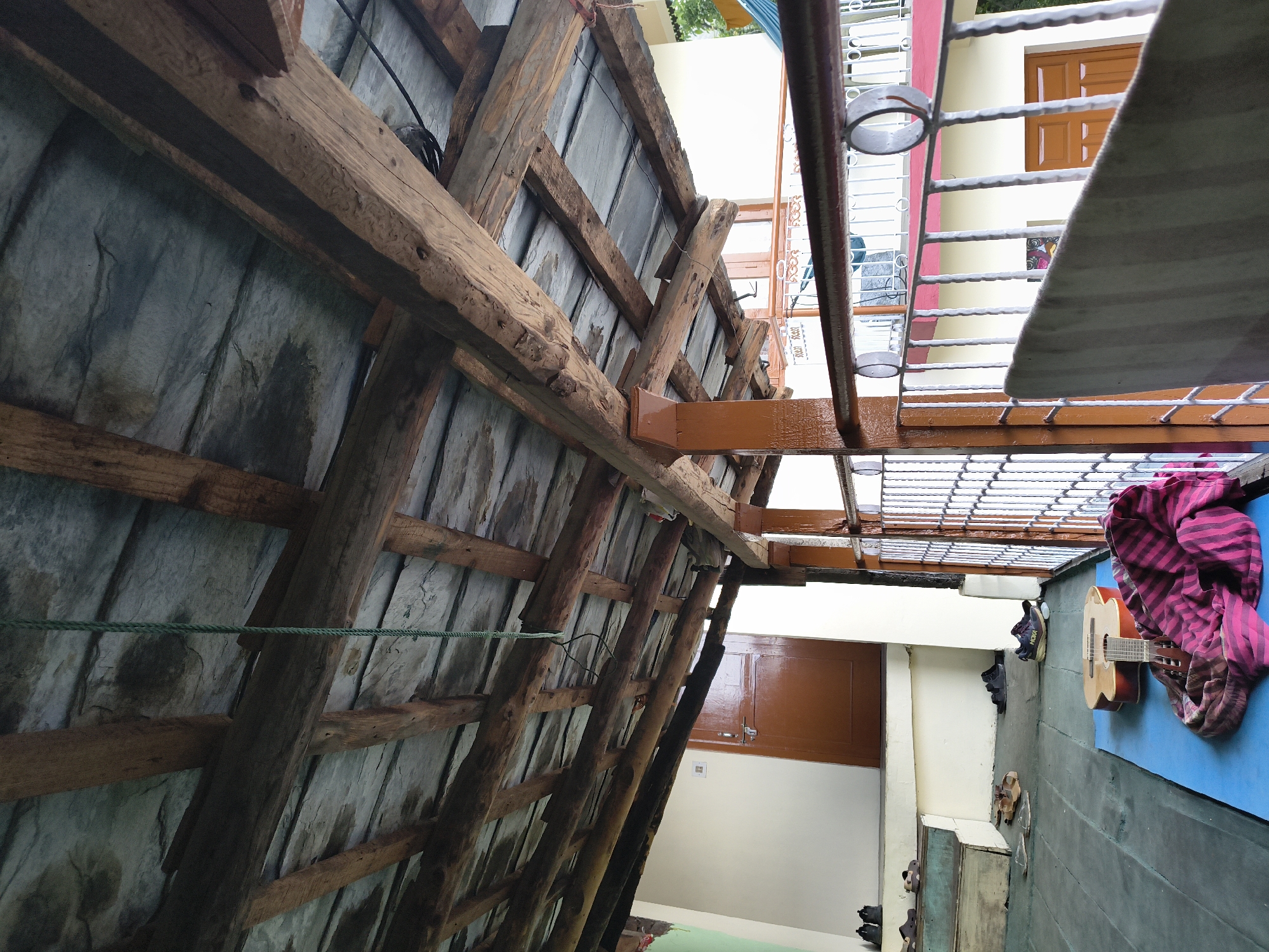 View from underneath the roof of a structure in Haini, India, looking out onto an open area. The roof's wooden framework is visible, supporting gray slate or tile. Outside the roof, there's a metal railing and the exterior wall of a building with a brown door. Below the railing and wall is a blue mat with a guitar and a striped pink cloth on top of it. Pairs of shoes are arranged on the ground near a set of stairs.